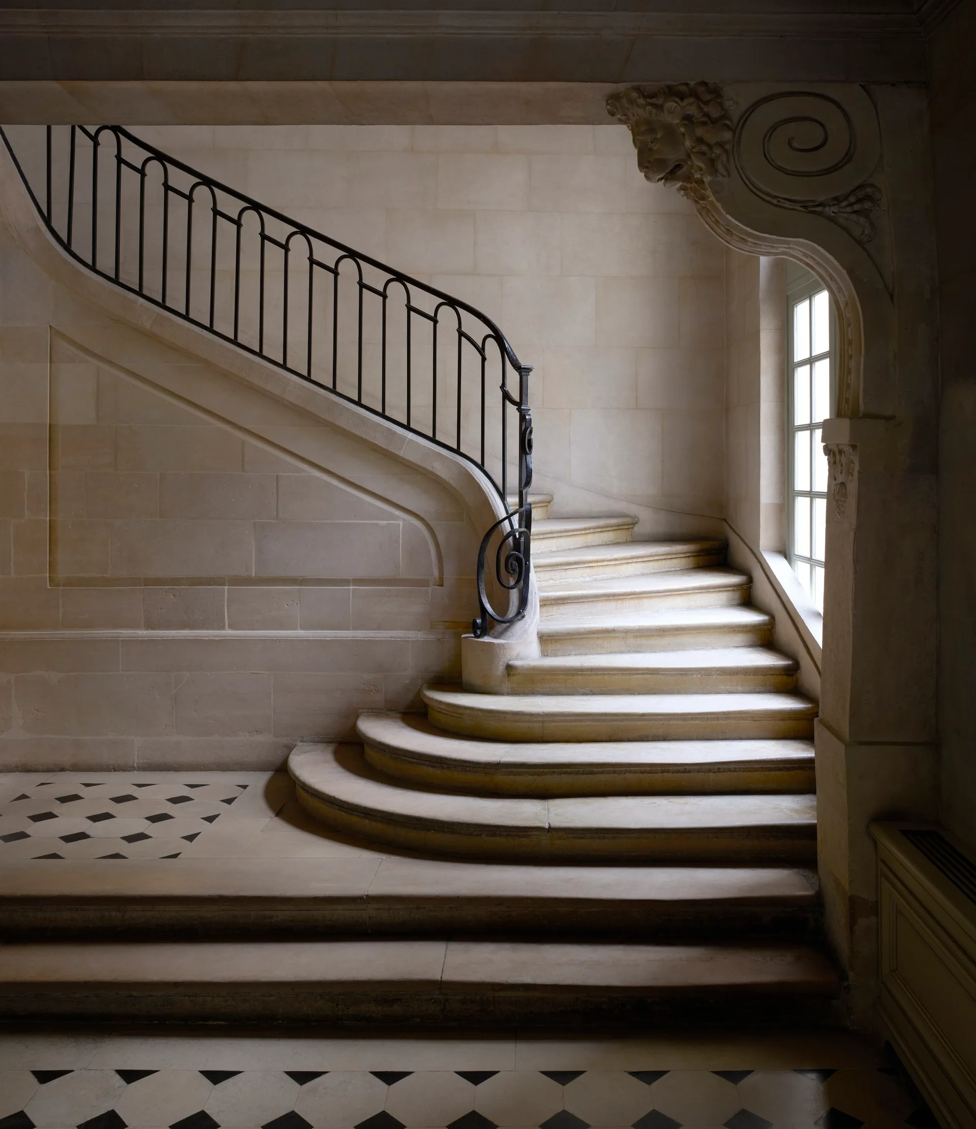 A curved beige stone staircase with a black wrought iron railing features a checkerboard tile floor and a carved stone archway.