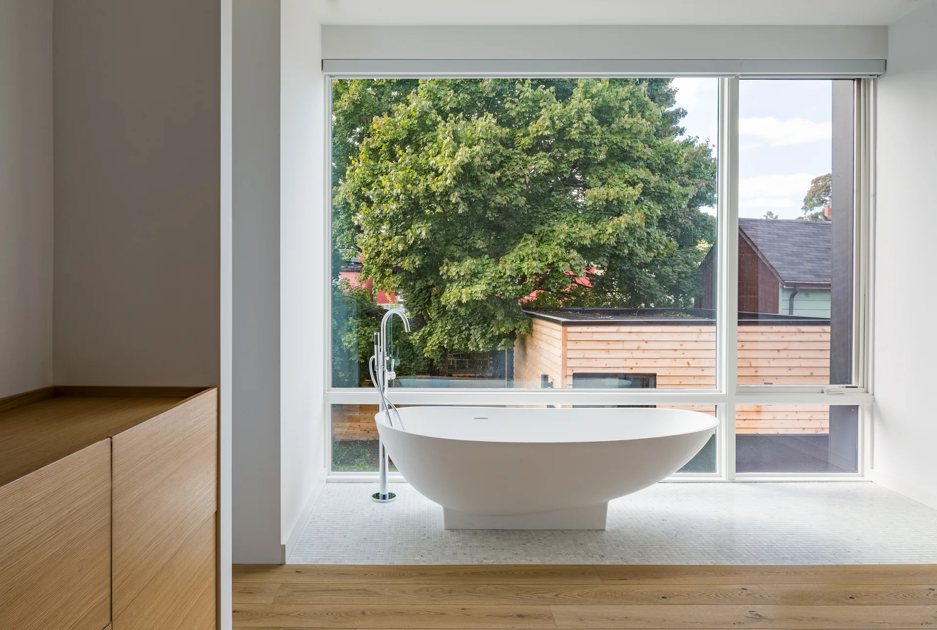 A minimalist bathroom features a white freestanding bathtub on a mosaic tile inset in front of a large picture window looking out at a green tree.