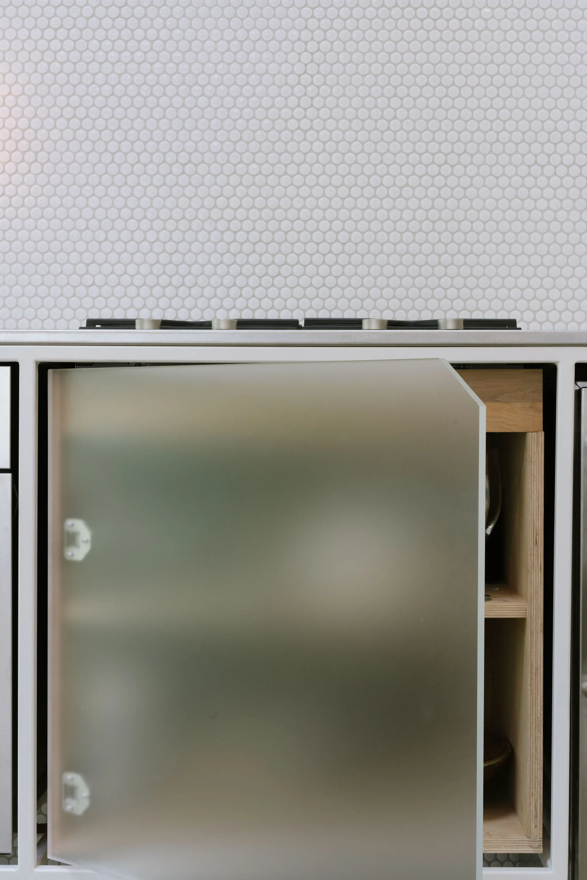 A modern kitchen cabinet features a semi-transparent frosted glass door revealing a light wood plywood interior set against a white penny tile backsplash.