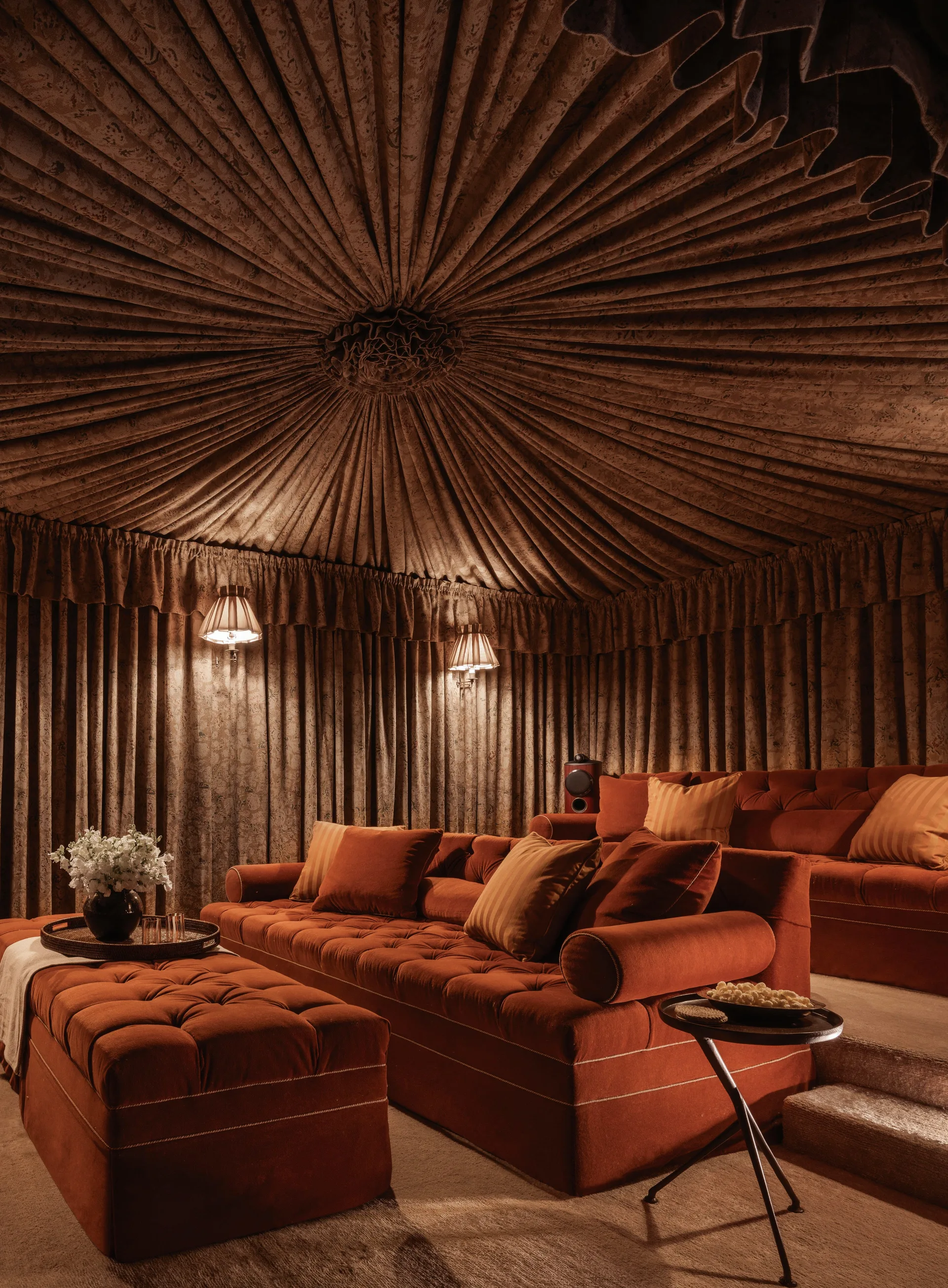 A theatrical home theater room featuring tiered terracotta velvet tufted sofas, a matching ottoman, and a pleated brown fabric-draped ceiling.