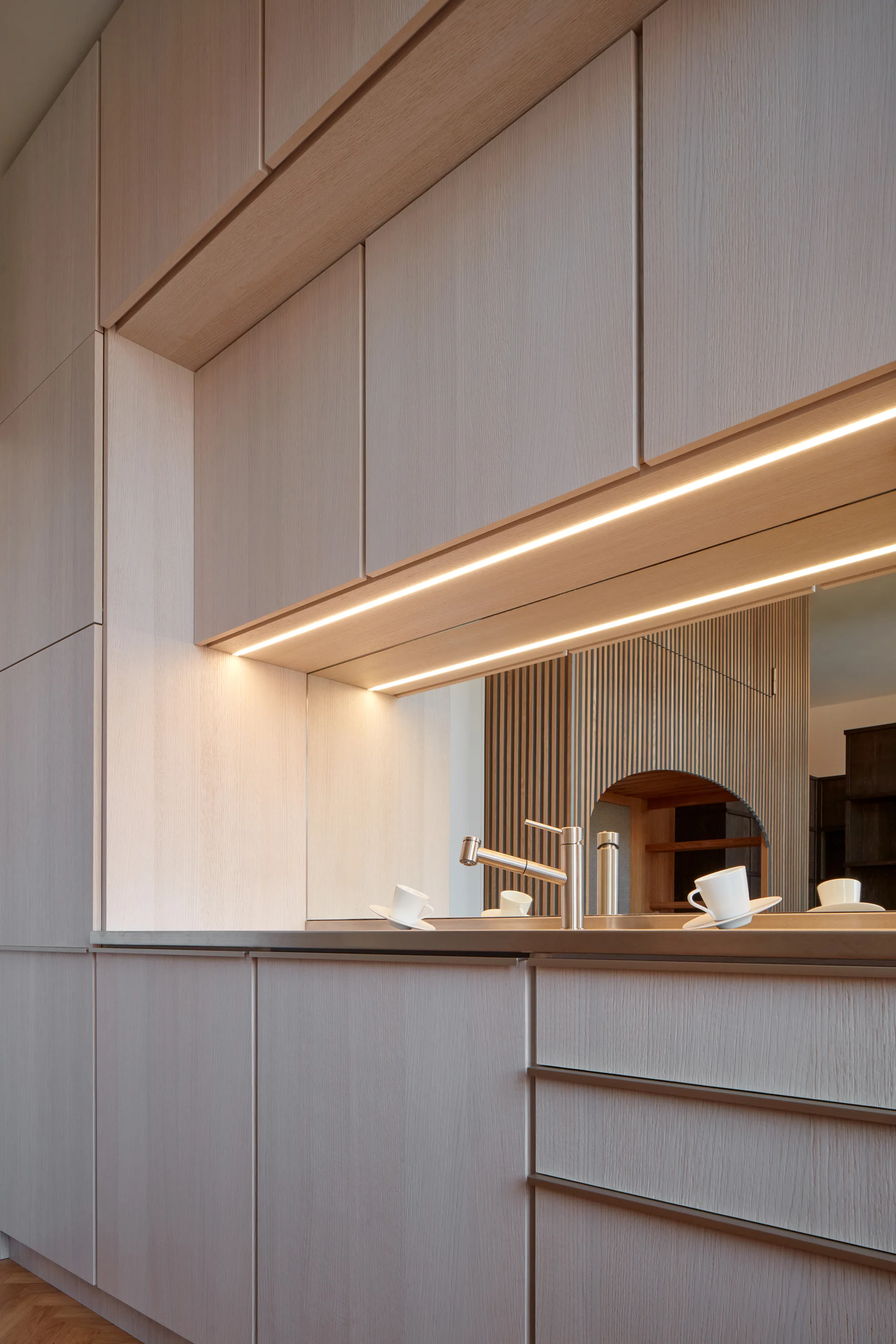 A close-up of light wood kitchen cabinetry featuring a mirrored backsplash, integrated LED lighting, and a modern steel faucet.