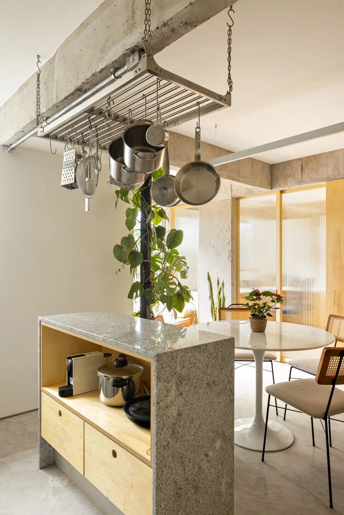 A granite-topped kitchen island with wood cabinetry stands beneath a suspended metal pot rack near a round dining table and ribbed glass doors.