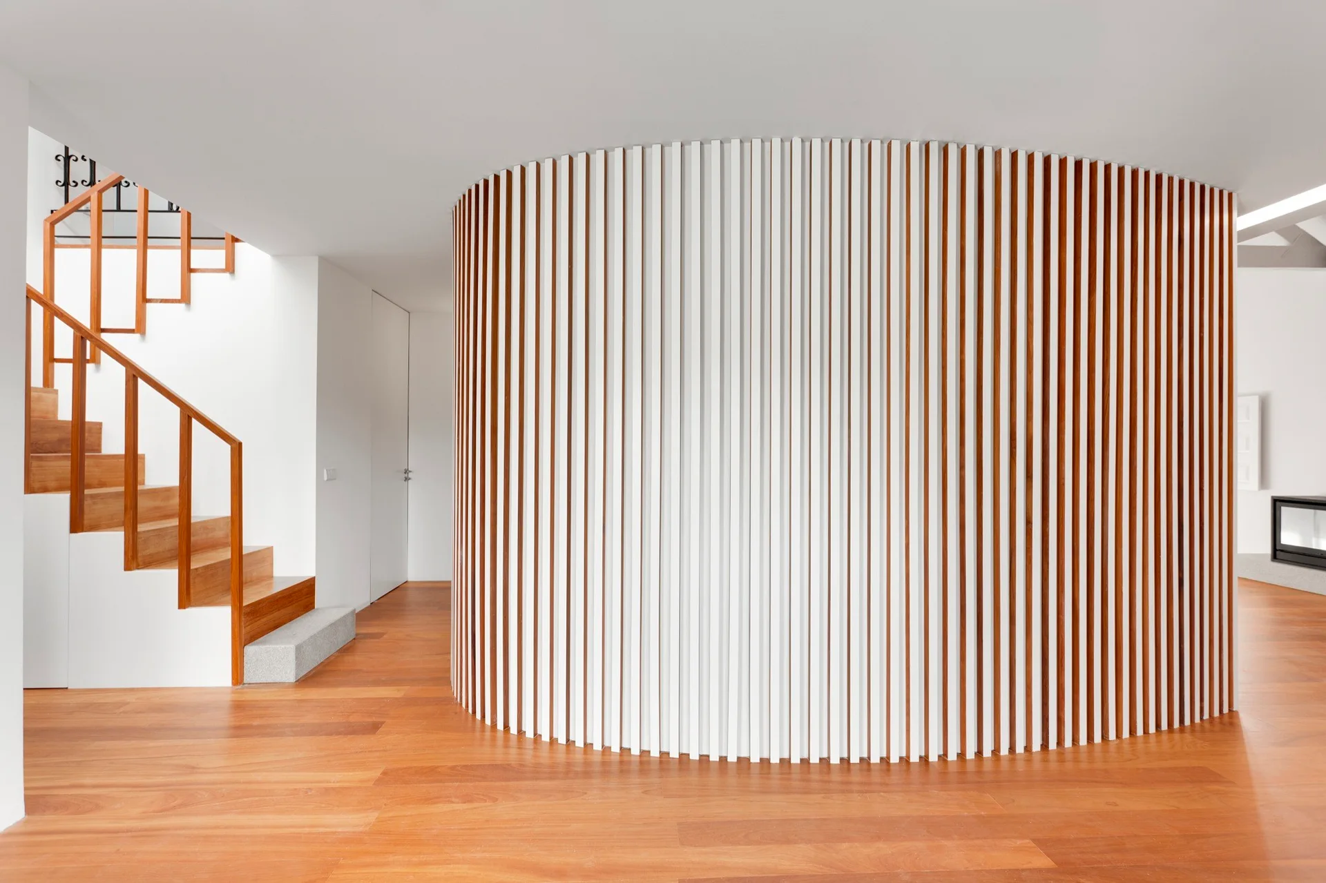 A modern hallway features a prominent curved wall with white and wood vertical slats, a wooden staircase, and warm hardwood flooring.