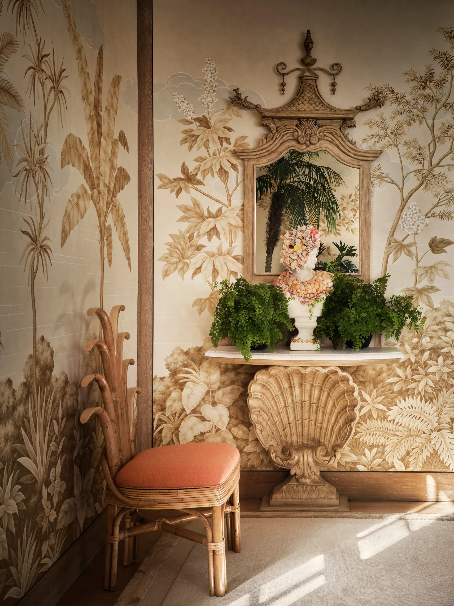 An entryway vignette features a shell-shaped carved console table, a bamboo chair with a coral cushion, and walls covered in a beige botanical mural.