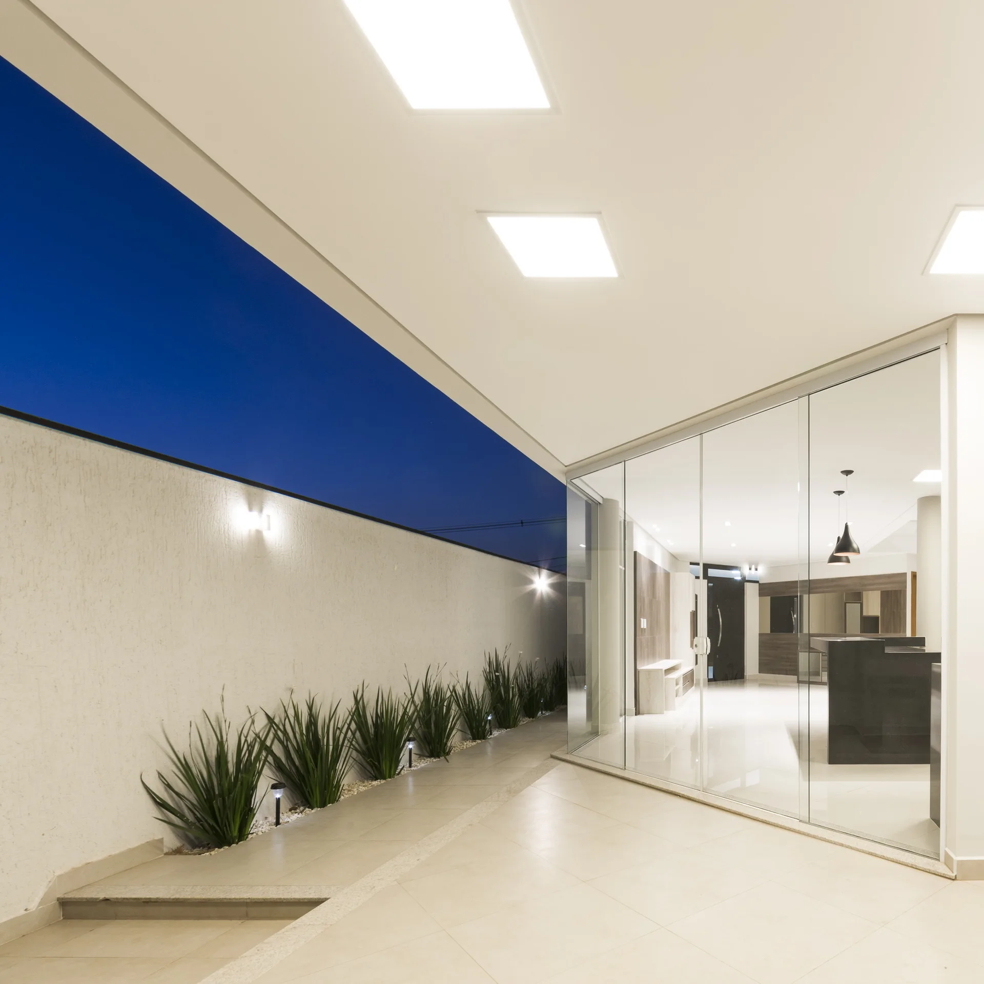 A modern covered patio features beige tiled flooring, square recessed ceiling lights, a glass partition wall, and a low landscaping bed along a plaster wall.