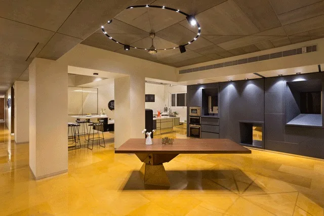 A modern dining and kitchen area features a dark wood cantilevered table set on a polished yellow stone floor beneath a circular ceiling light.