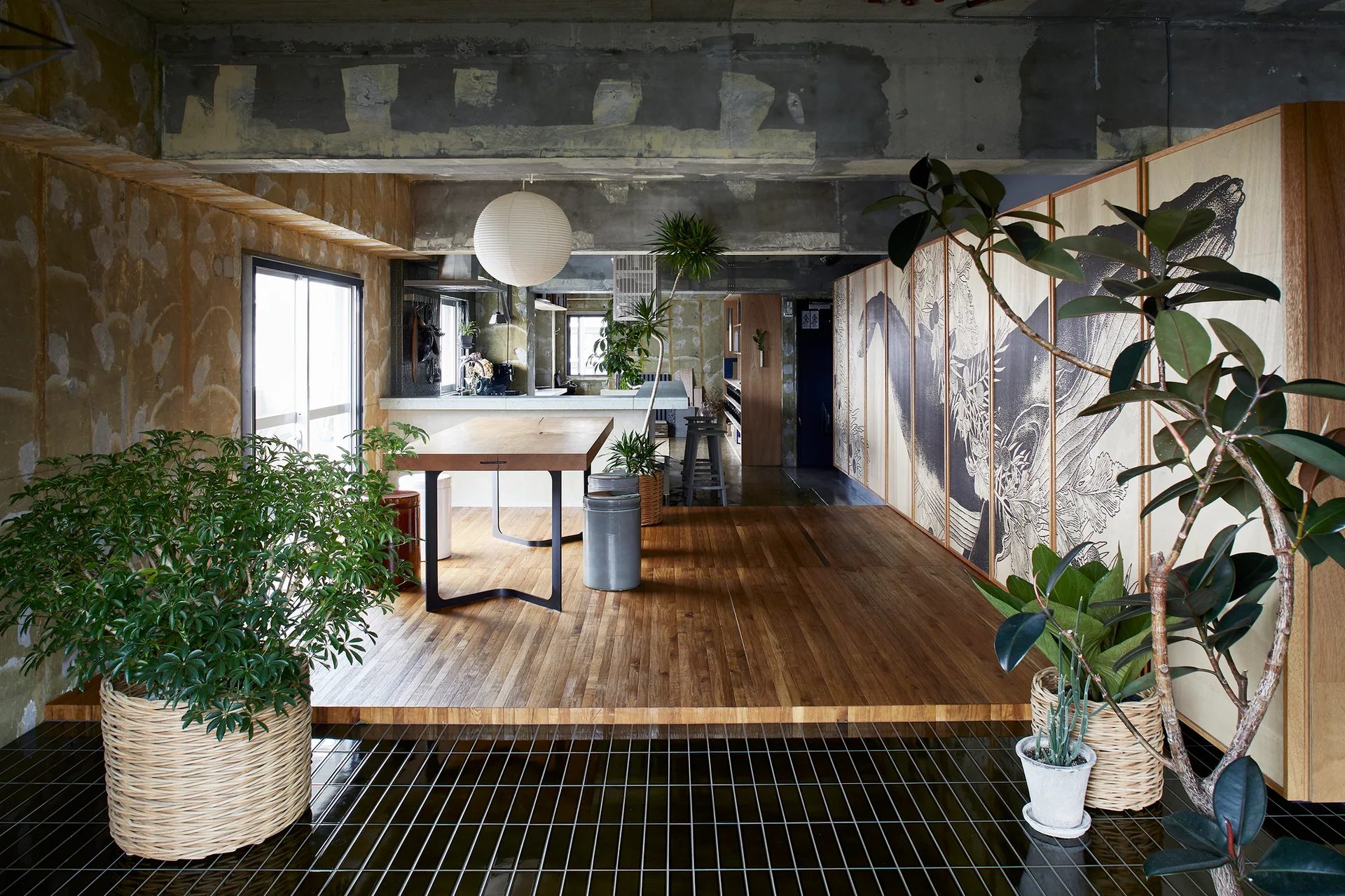 An industrial open-plan dining space with a raised wooden platform, exposed concrete walls, a round paper pendant, and large graphic art cabinets.