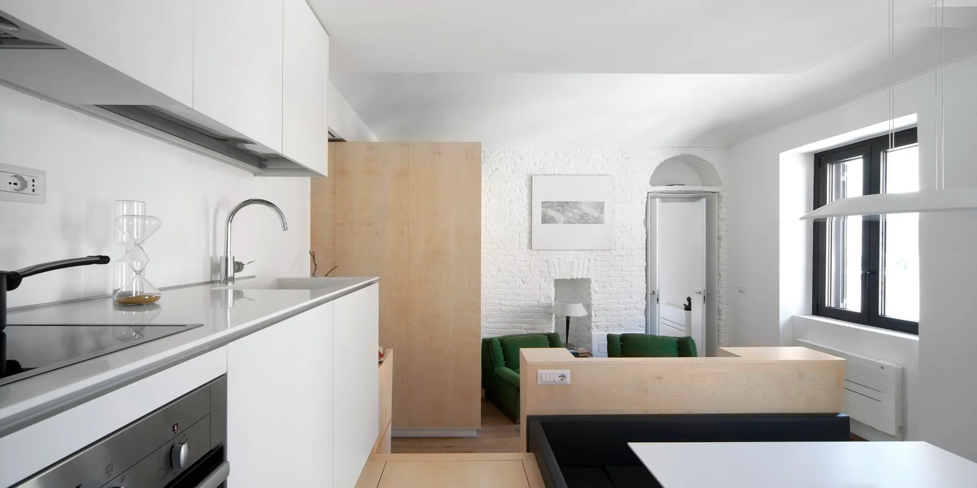 A modern open-plan kitchen with white handleless cabinets and light wood partitions leading to a living area with two green chairs and a white brick wall.
