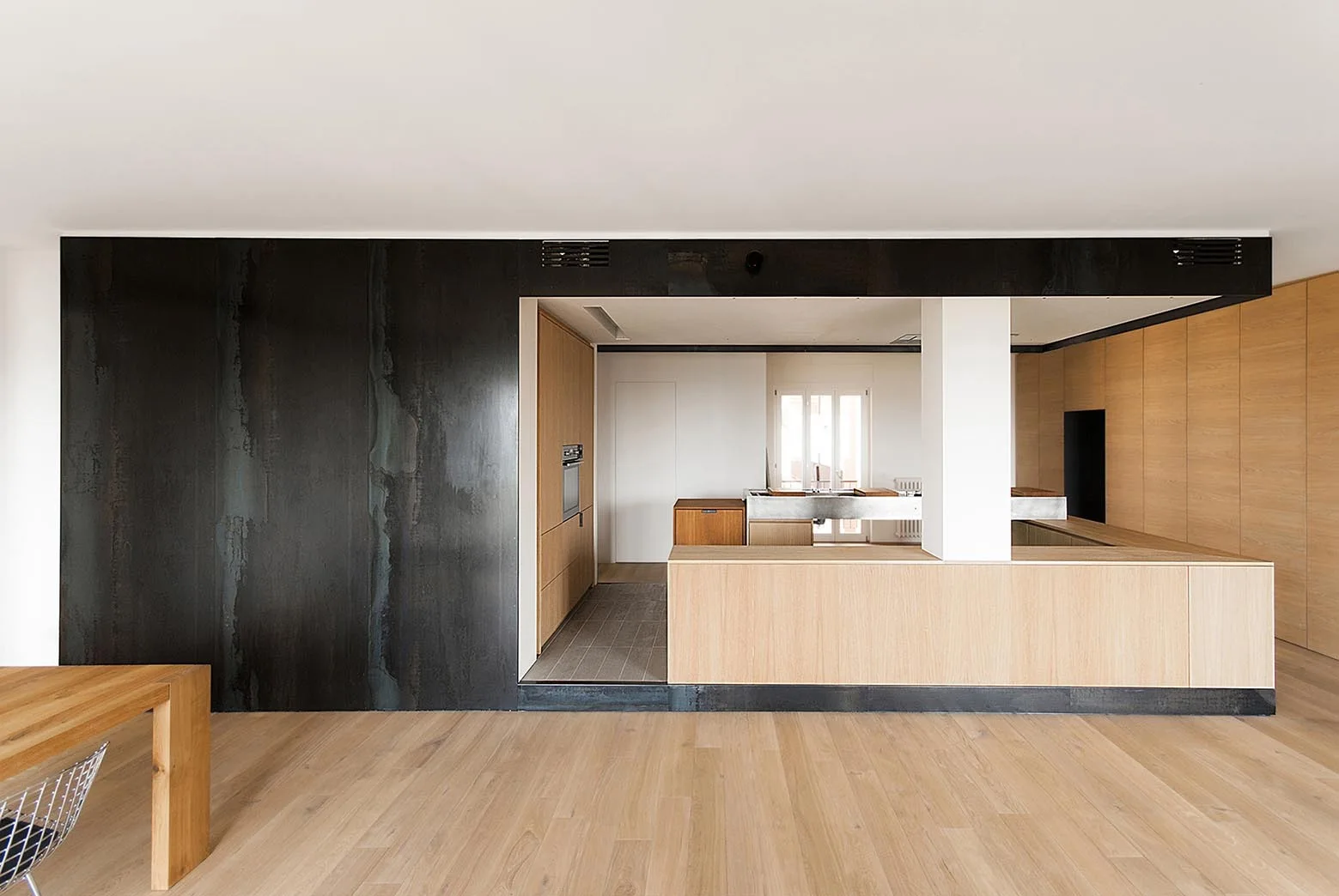 A modern open-plan kitchen features a large dark metal wall panel, light wood cabinetry, a central island, and pale wood flooring.