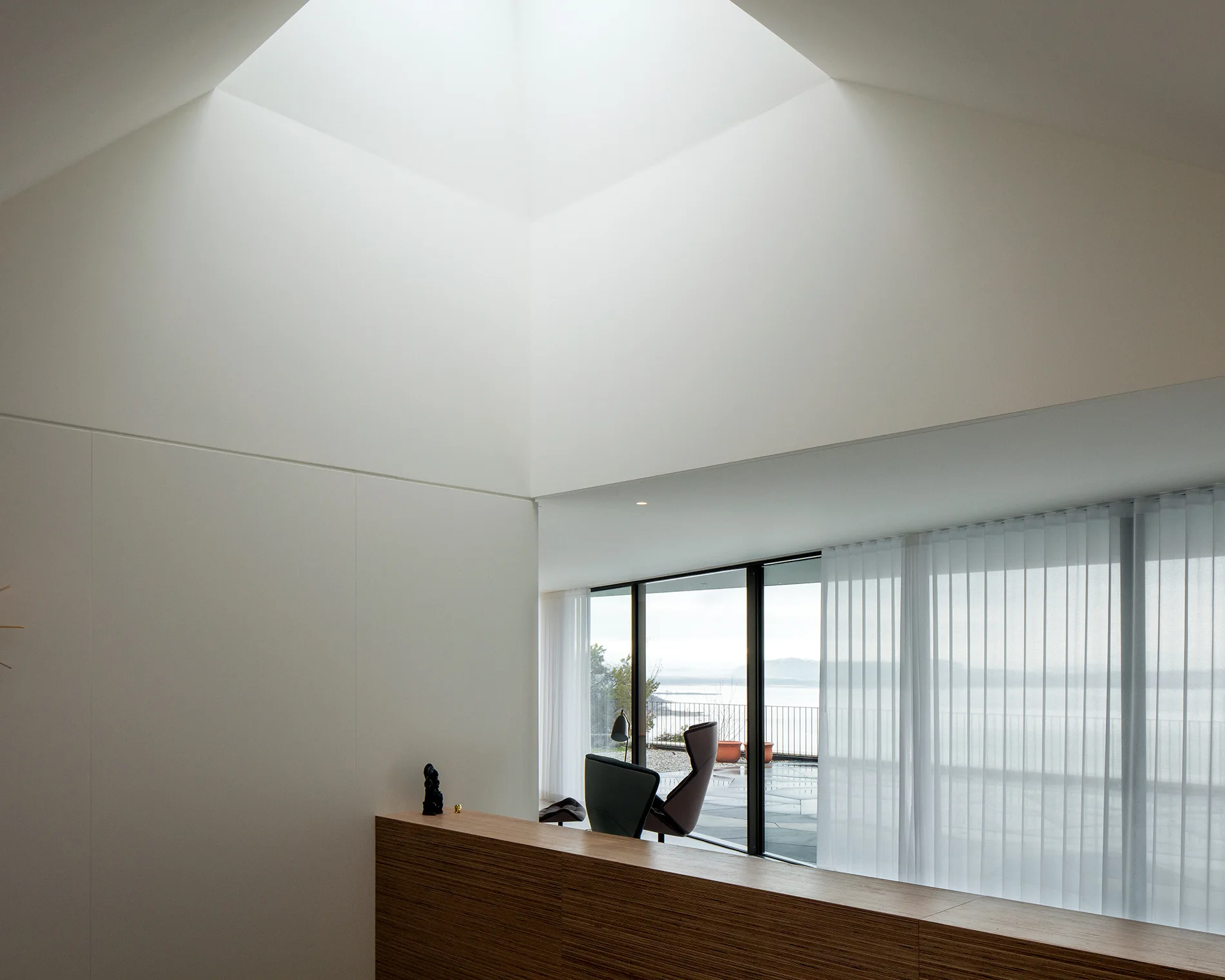 A minimalist white living area features a large geometric ceiling skylight, a wood-toned console, and a glass wall with sheer curtains in the background.
