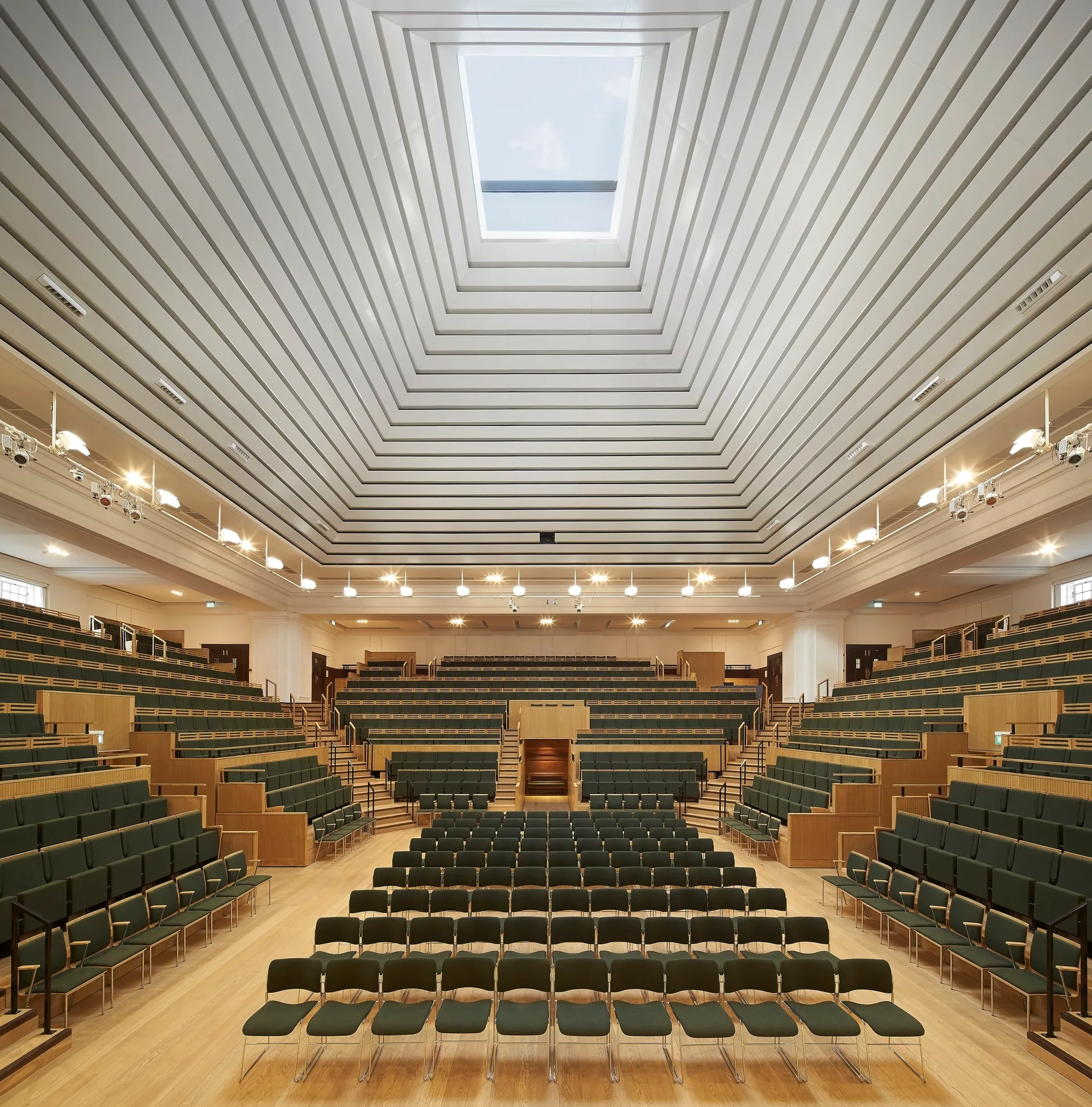 A large lecture hall featuring tiers of dark green seating under a white stepped ceiling with a large central skylight.