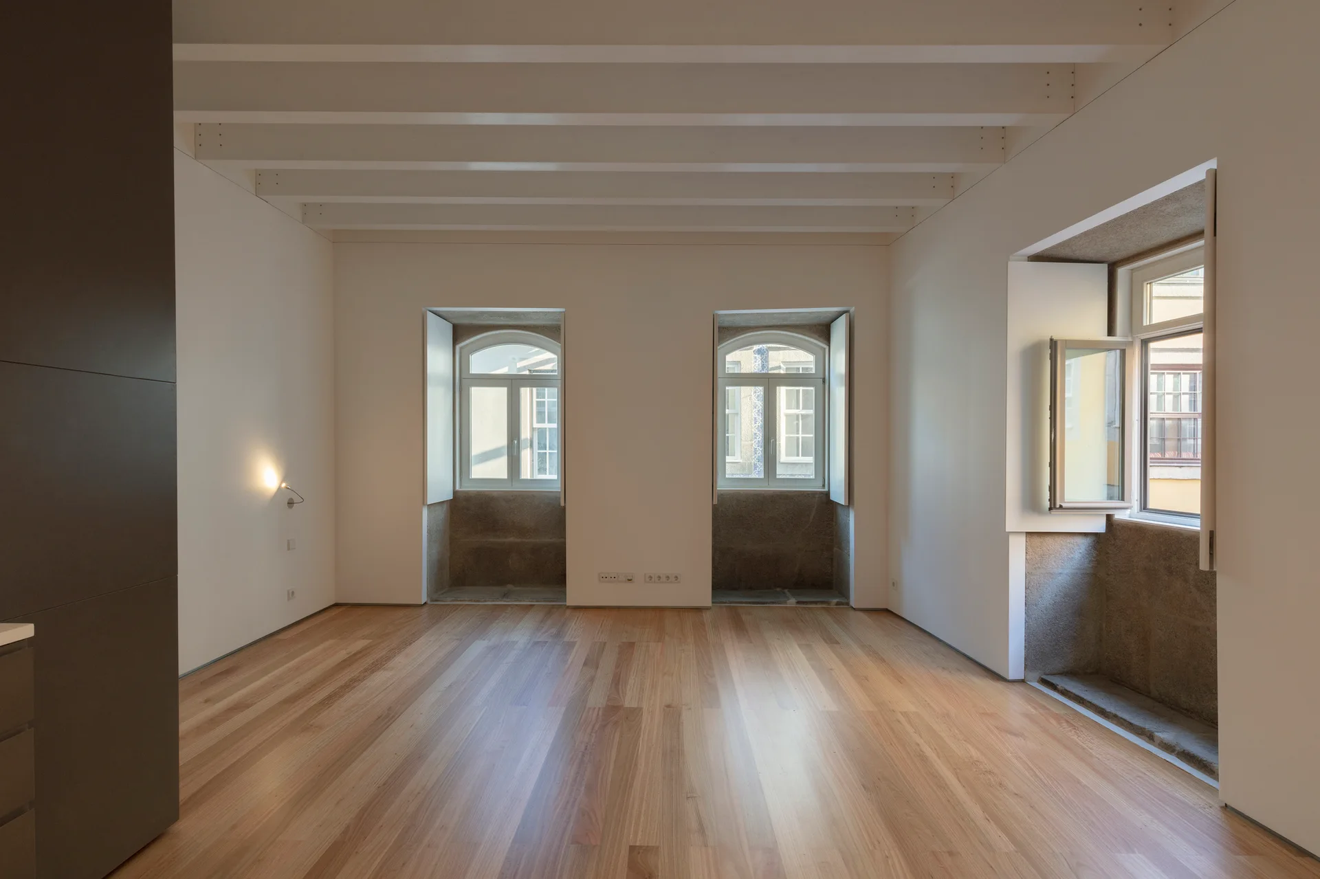 An empty room features light wood flooring, white walls with exposed ceiling beams, and three deep recessed windows with stone sills.