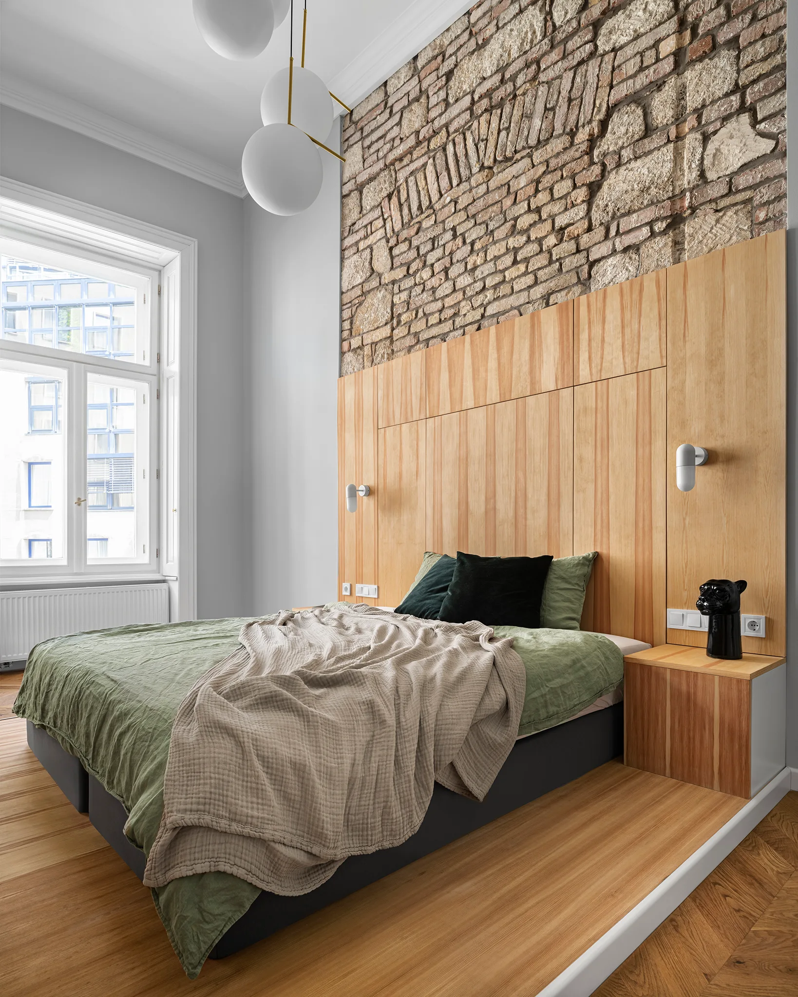 A modern bedroom features an integrated wood headboard set against an exposed brick wall, paired with a green-dressed bed and spherical pendant lights.