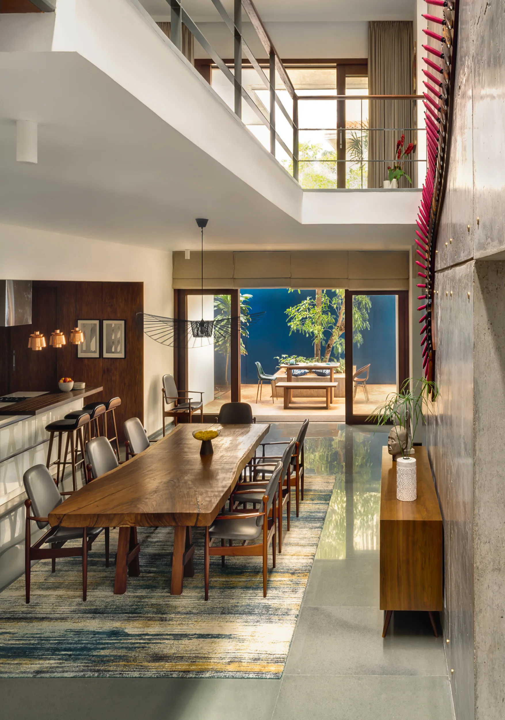 An open-plan dining room features a large live-edge wood table, mid-century chairs, and an exposed concrete wall under a mezzanine balcony.