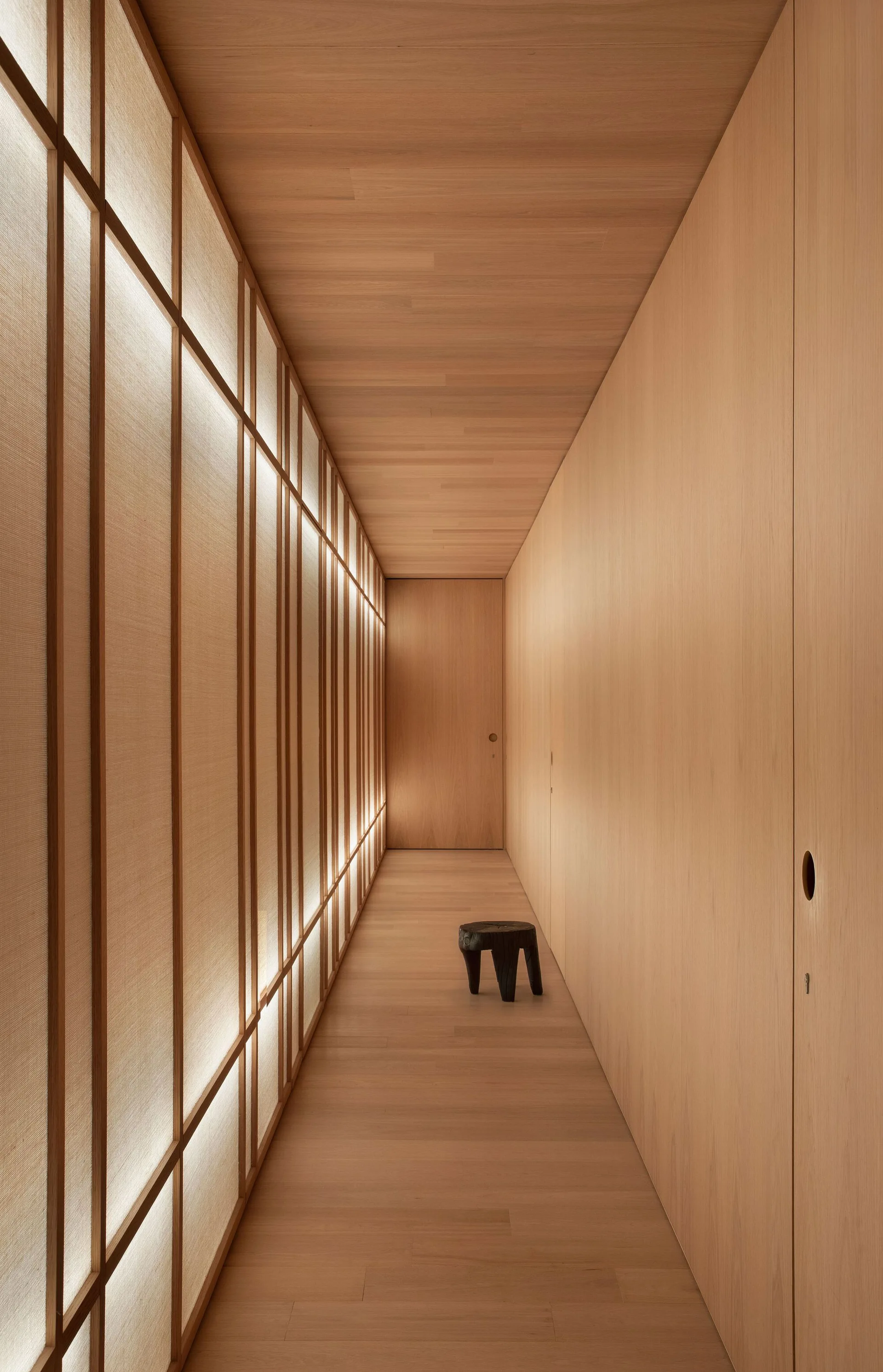 A minimalist wood-paneled hallway featuring a warm backlit grid screen wall, integrated flush doors, and a small black wood stool.