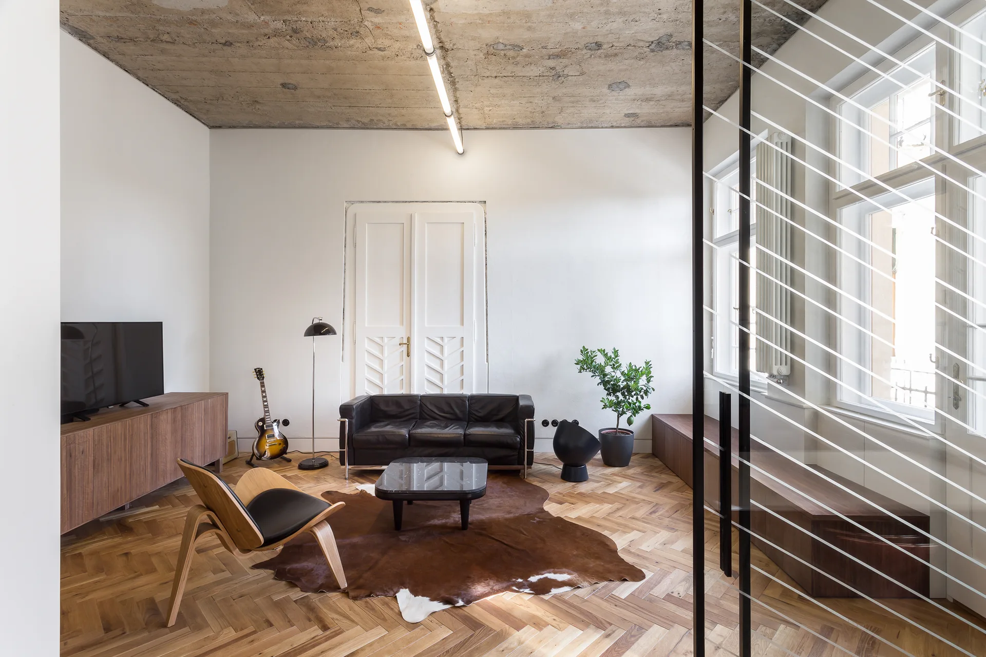 A modern minimalist living room features a black leather sofa and a cowhide rug on herringbone floors under a concrete ceiling.
