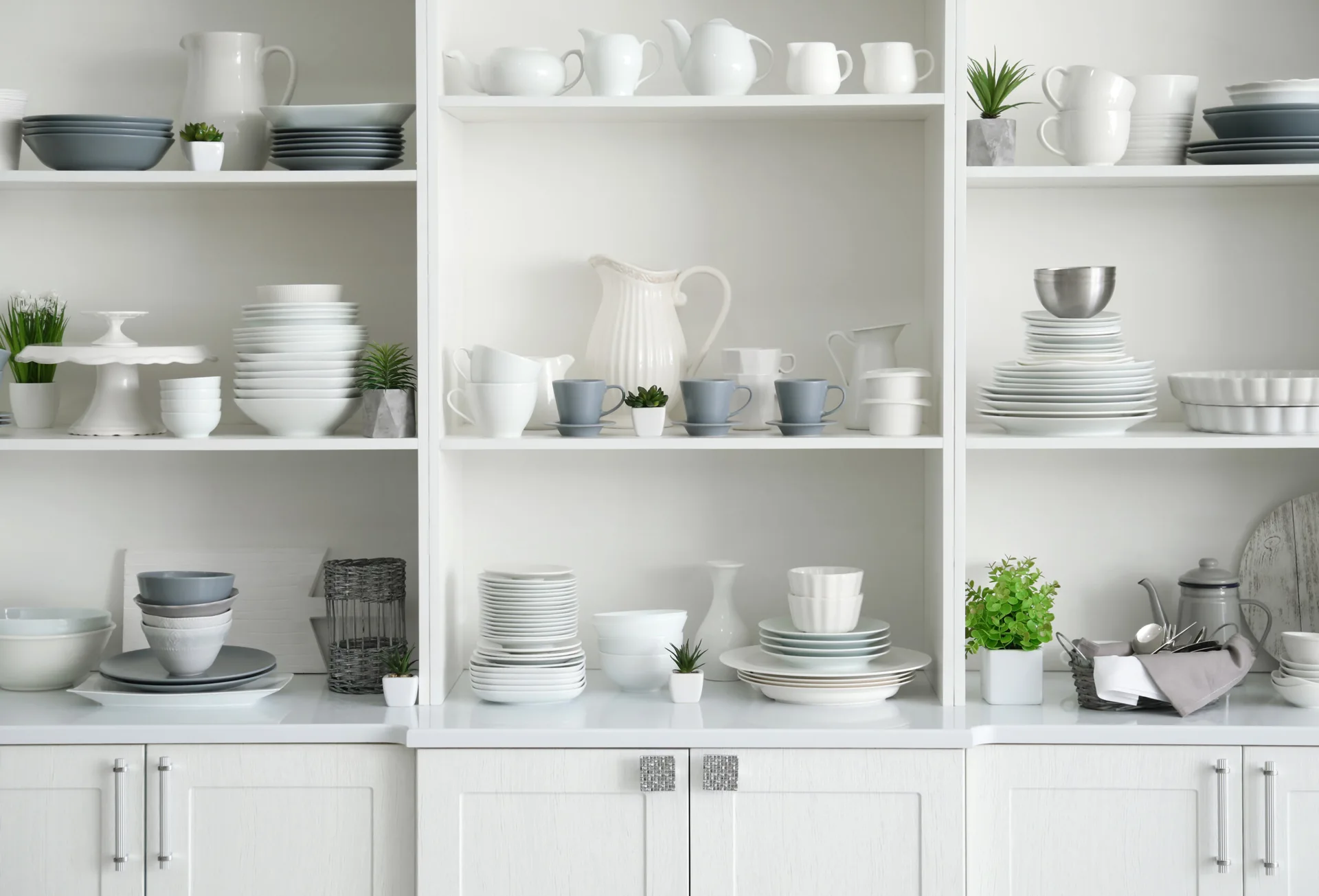 White built-in kitchen cabinet shelves displaying neatly arranged stacks of grey and white ceramic plates, bowls, mugs, and small potted plants.