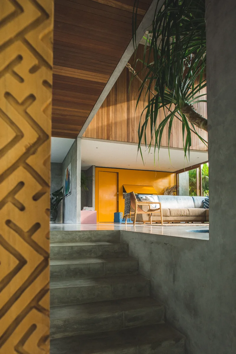 A concrete staircase leads up to a modern open-plan living room with a wood ceiling and a bold yellow accent wall.