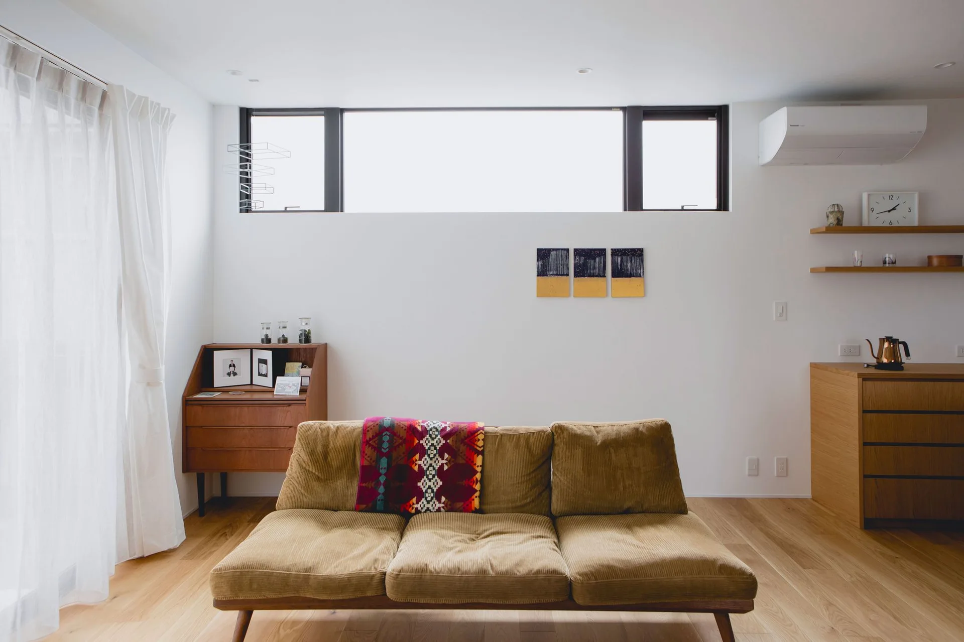 A minimalist living room features a tan corduroy sofa, wooden cabinets, and a high horizontal window against plain white walls.