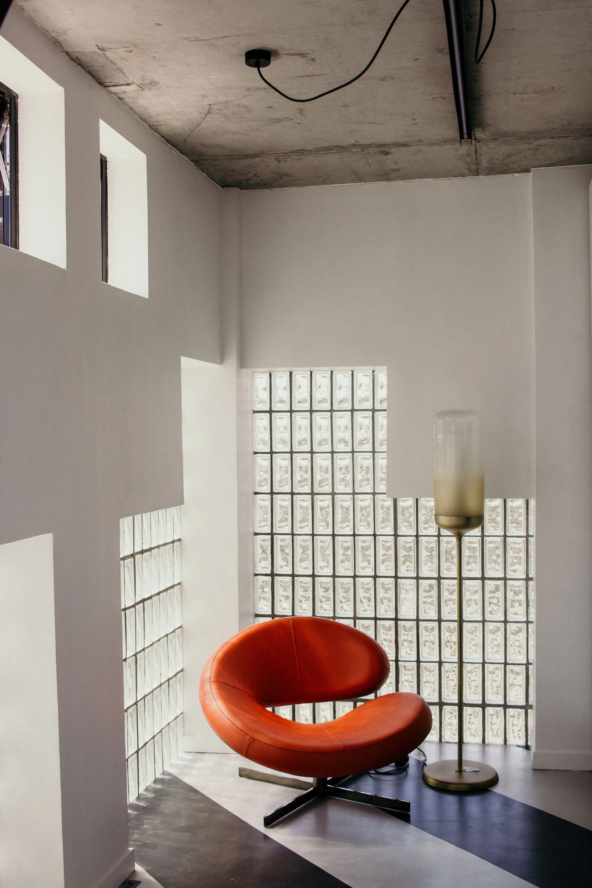 A curved orange swivel armchair sits in a white room with a glass block wall, concrete ceiling, and tall brass floor lamp.