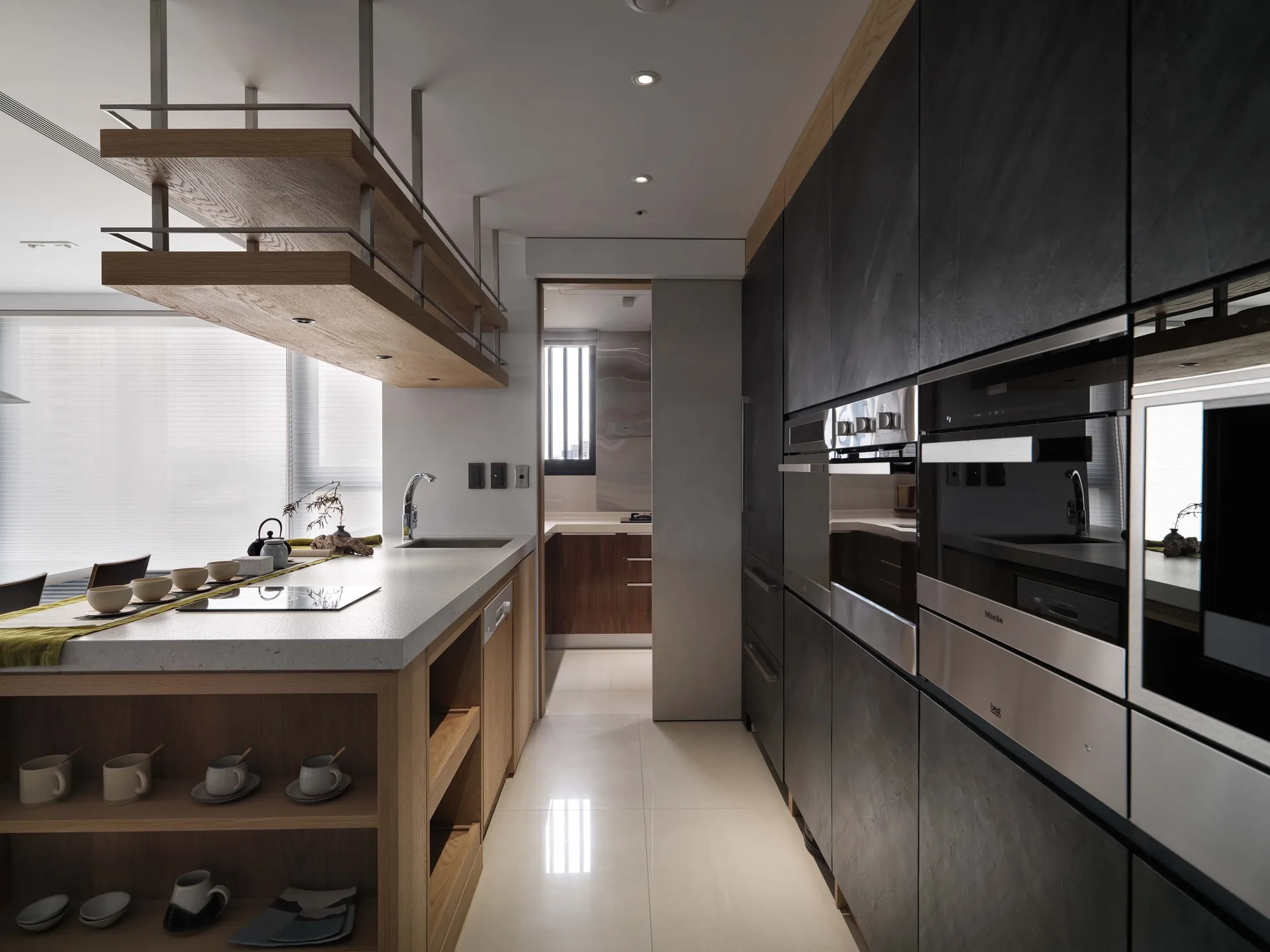 A modern kitchen featuring a dark charcoal cabinet wall with built-in ovens, a stone-topped island with wood shelving, and a suspended wooden ceiling rack.