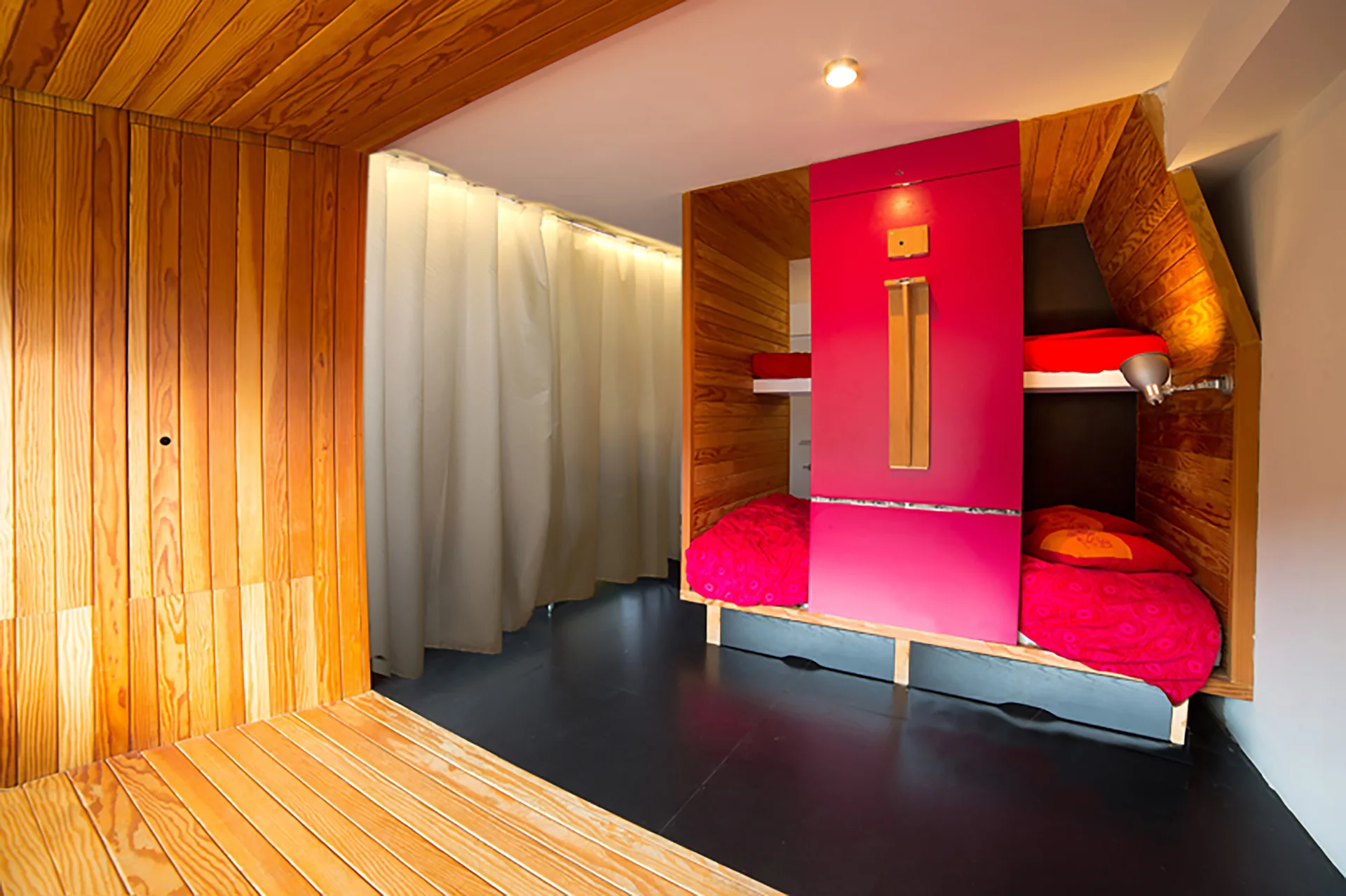 A wood-paneled sleeping alcove featuring a built-in bunk bed unit with a central pinkish-red panel, red bedding, and a black floor.
