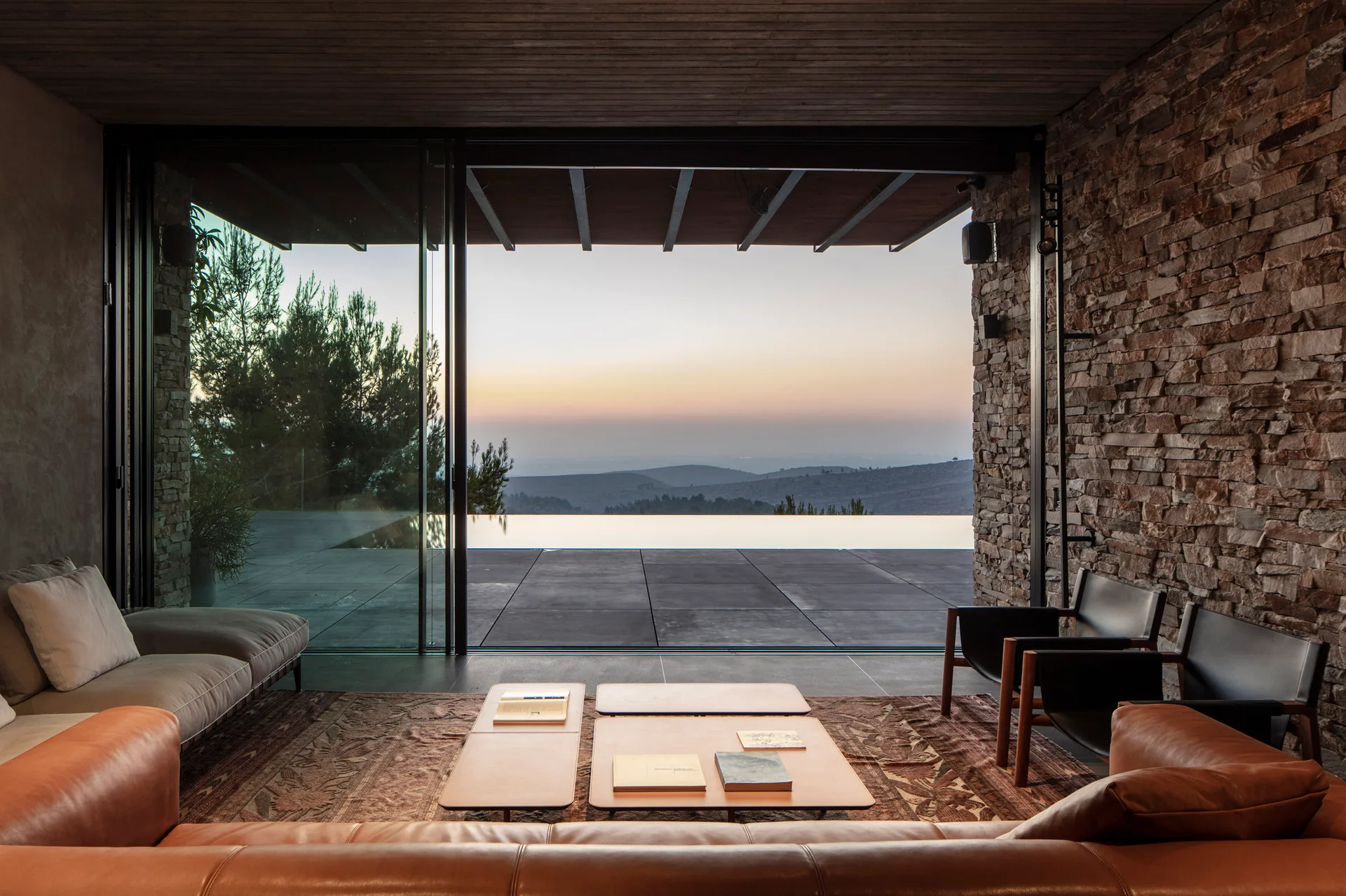 A modern living room with a tan leather sofa, stone wall, and large sliding glass doors opening to an outdoor patio and infinity pool at sunset.