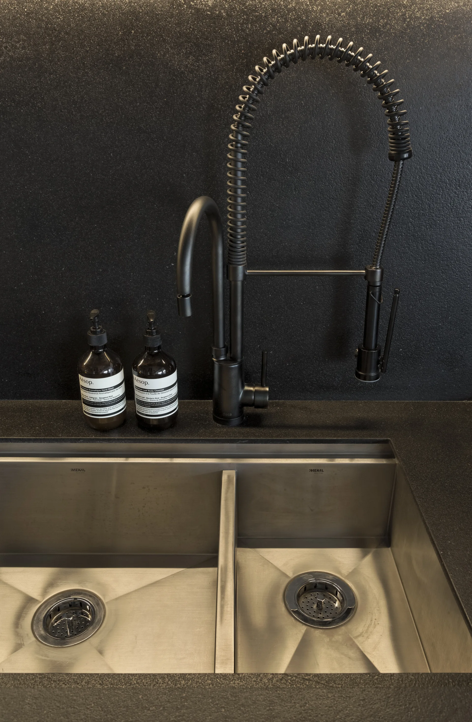A close-up of a matte black spring-neck faucet mounted on a dark stone countertop above a stainless steel double-basin kitchen sink.