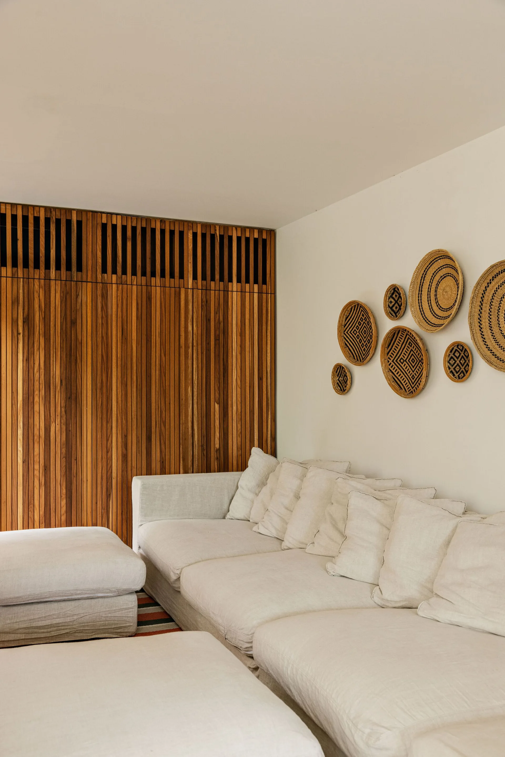 A large off-white linen modular sofa sits against a white wall decorated with woven baskets next to a vertical wood slat accent wall.