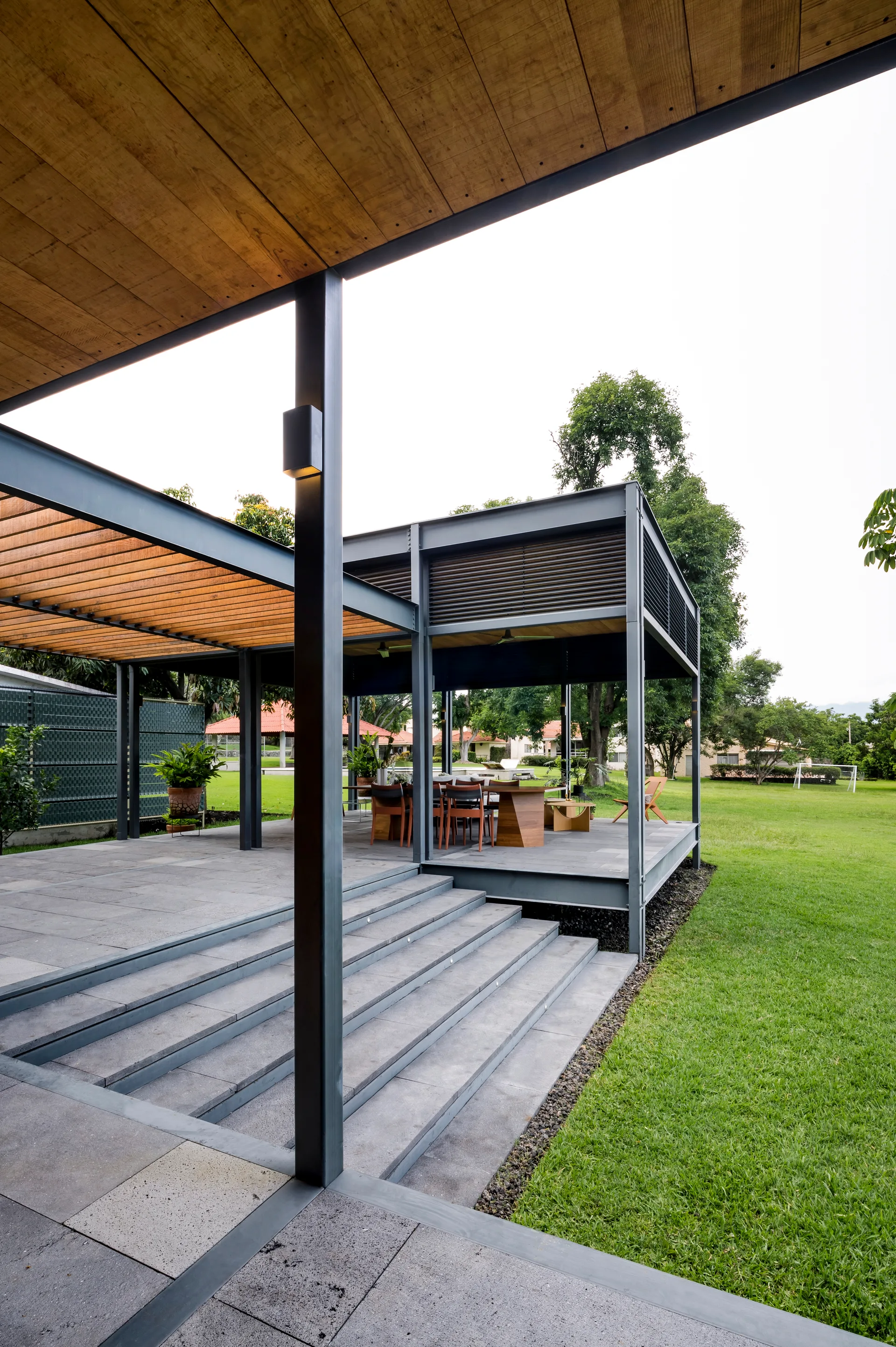 A modern covered outdoor patio featuring black metal framing, a wood-paneled ceiling, stone steps, and an outdoor dining table next to a grassy lawn.