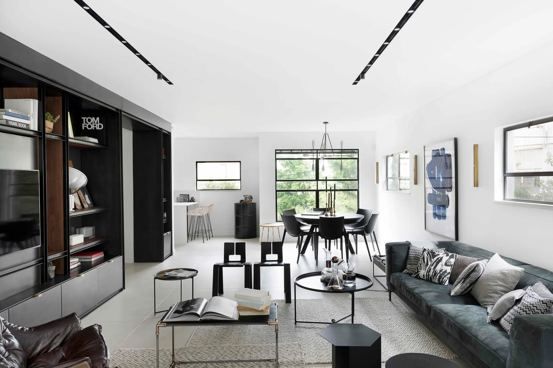 A modern open living room featuring a dark sofa, black nested coffee tables, a large built-in shelving unit, and a black dining set in the background.