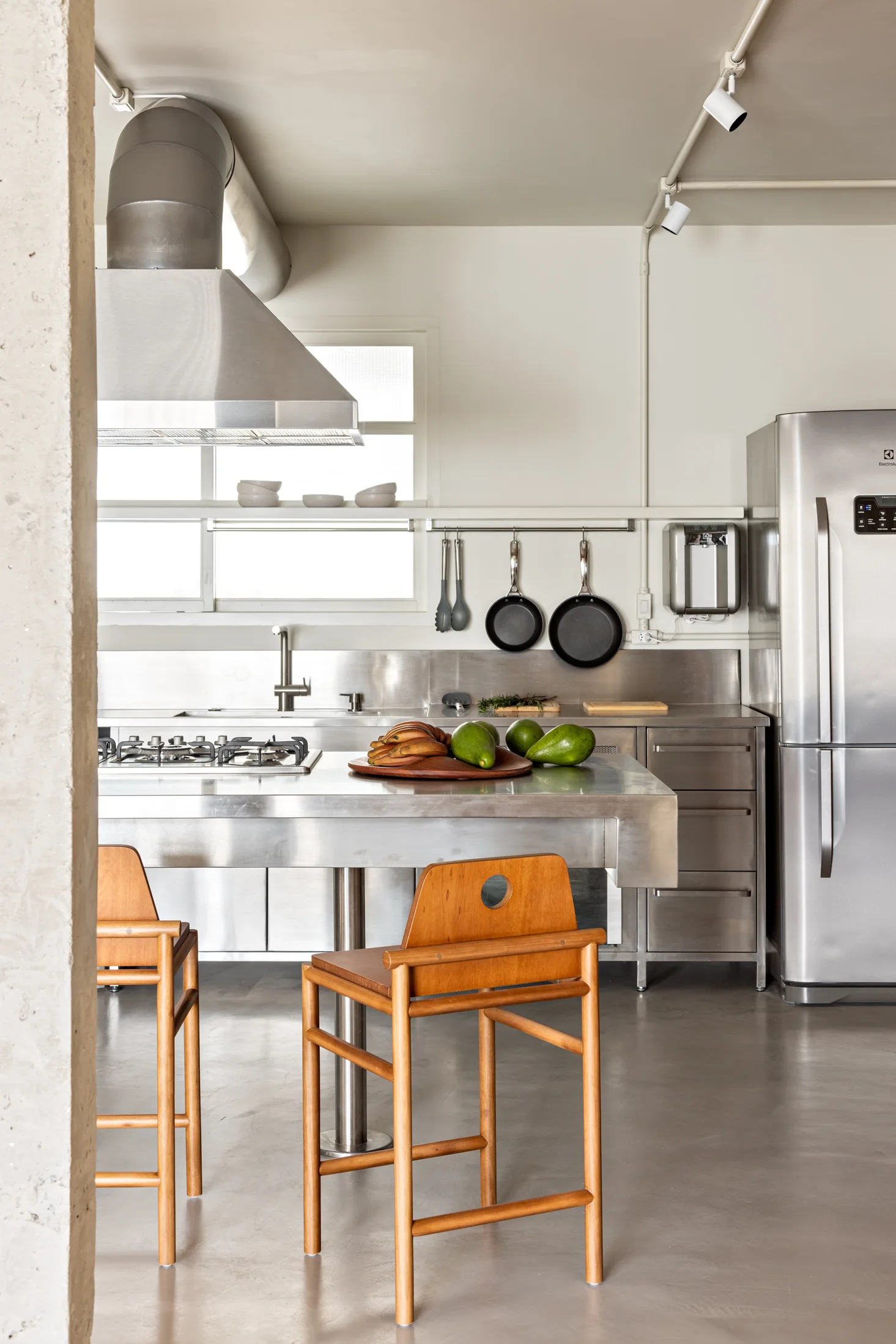 An industrial kitchen features a stainless steel island with wooden bar stools, a concrete floor, and matching metal cabinets and appliances.