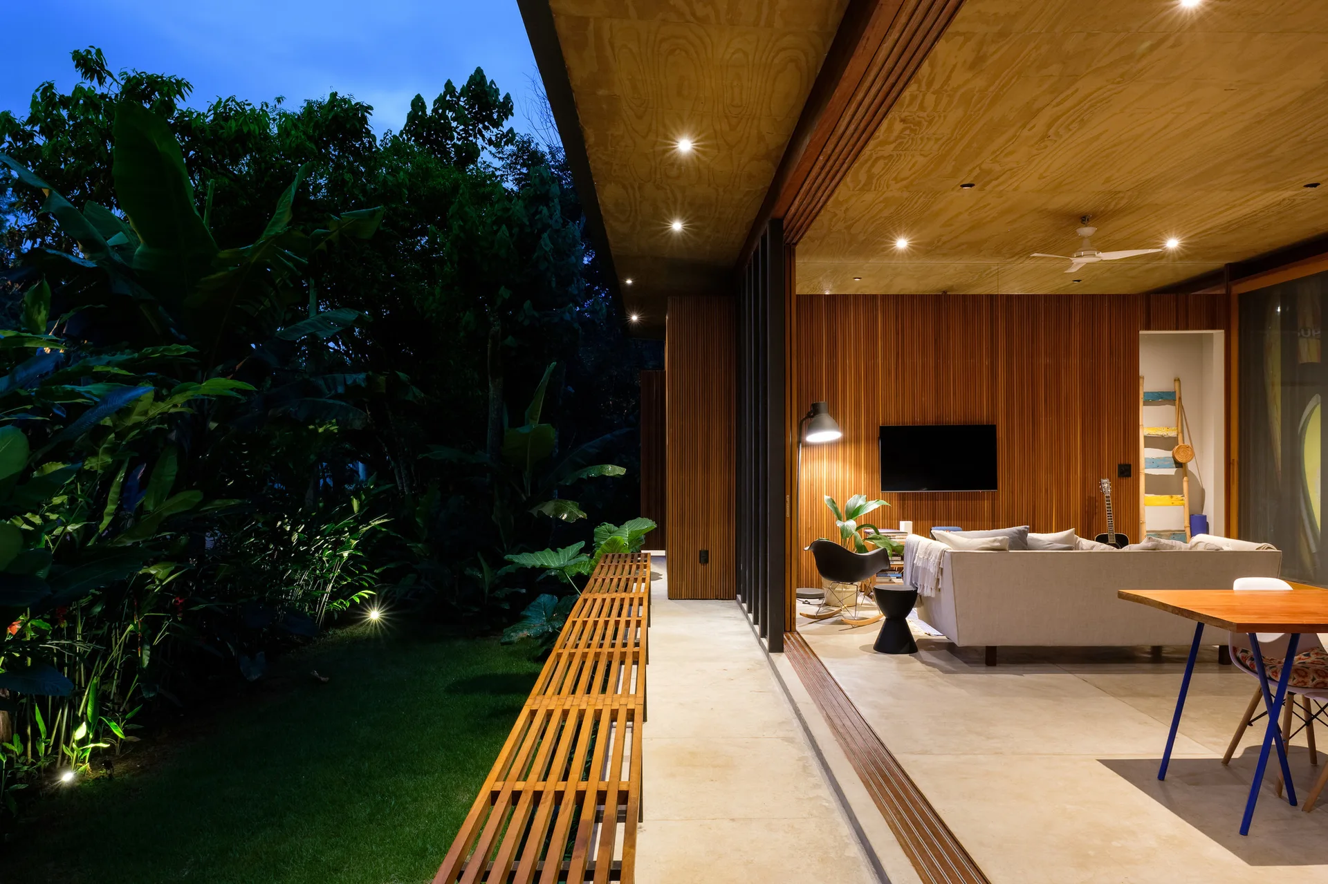 A modern living room with wood slat walls and a beige sofa opens onto a stone tile patio and an illuminated garden at night.