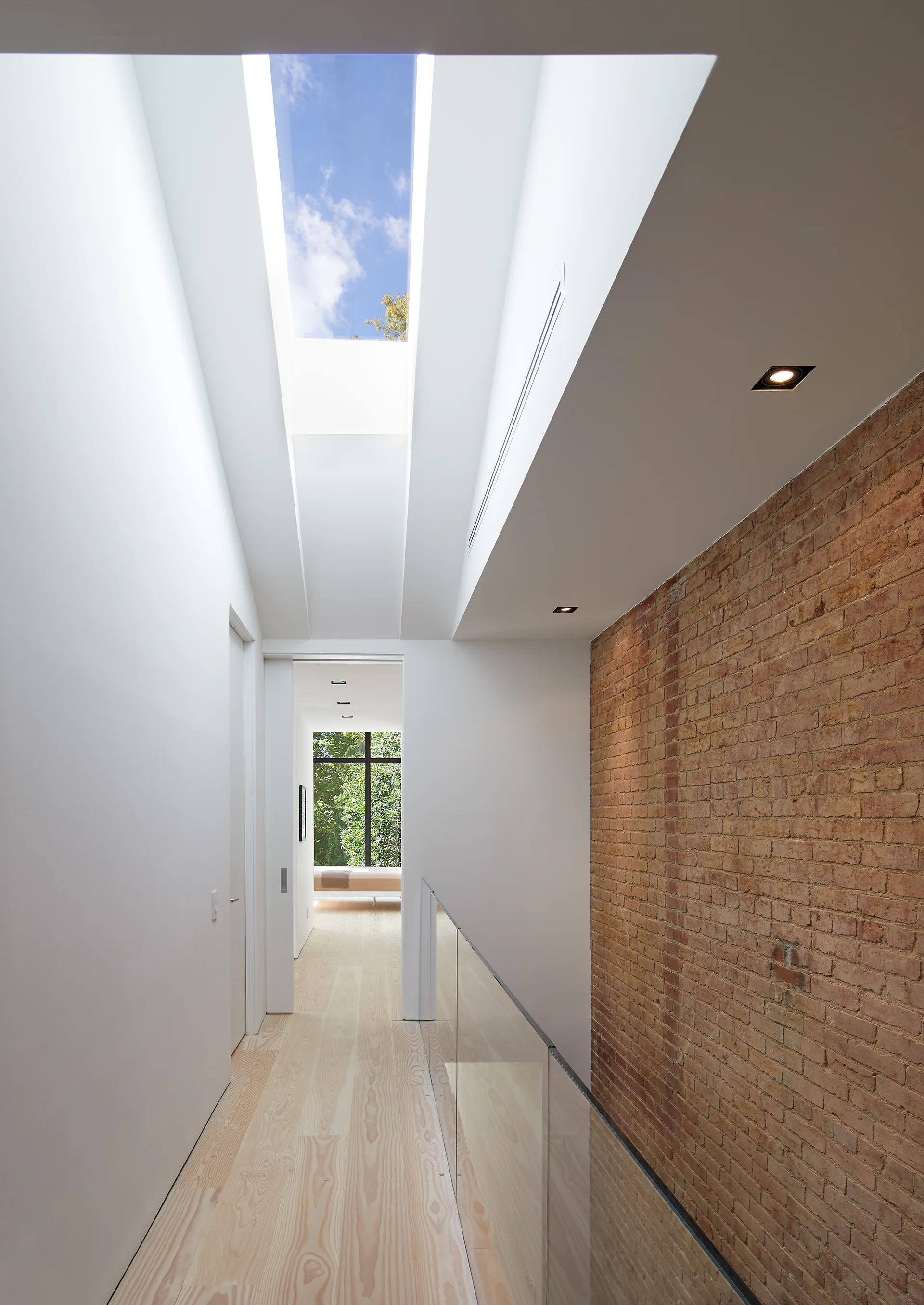 A bright modern hallway featuring a long skylight, an exposed red brick wall, light wood floors, and a glass railing.
