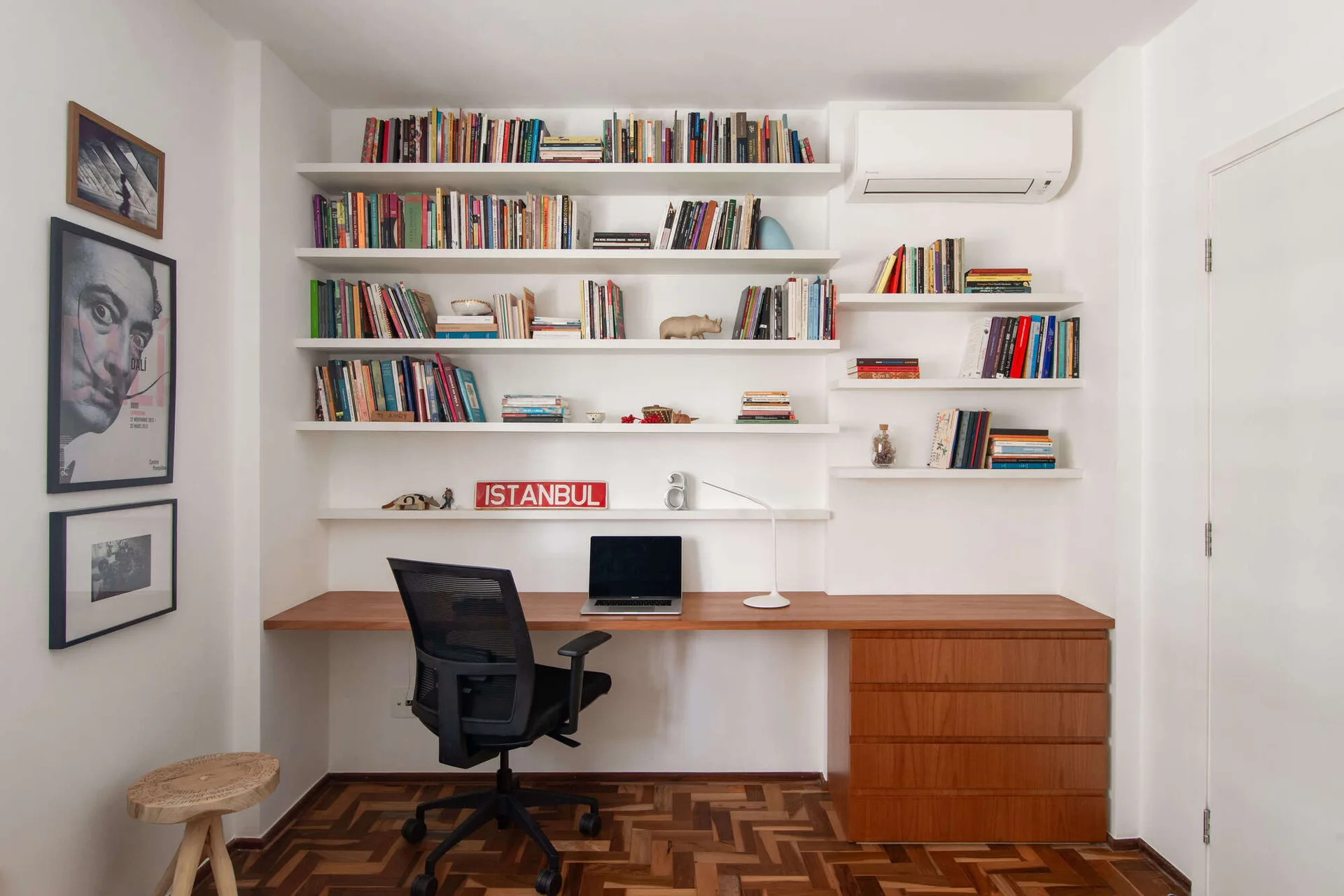A home office features a built-in wood desk, a black mesh office chair, and white shelves filled with books against white walls.