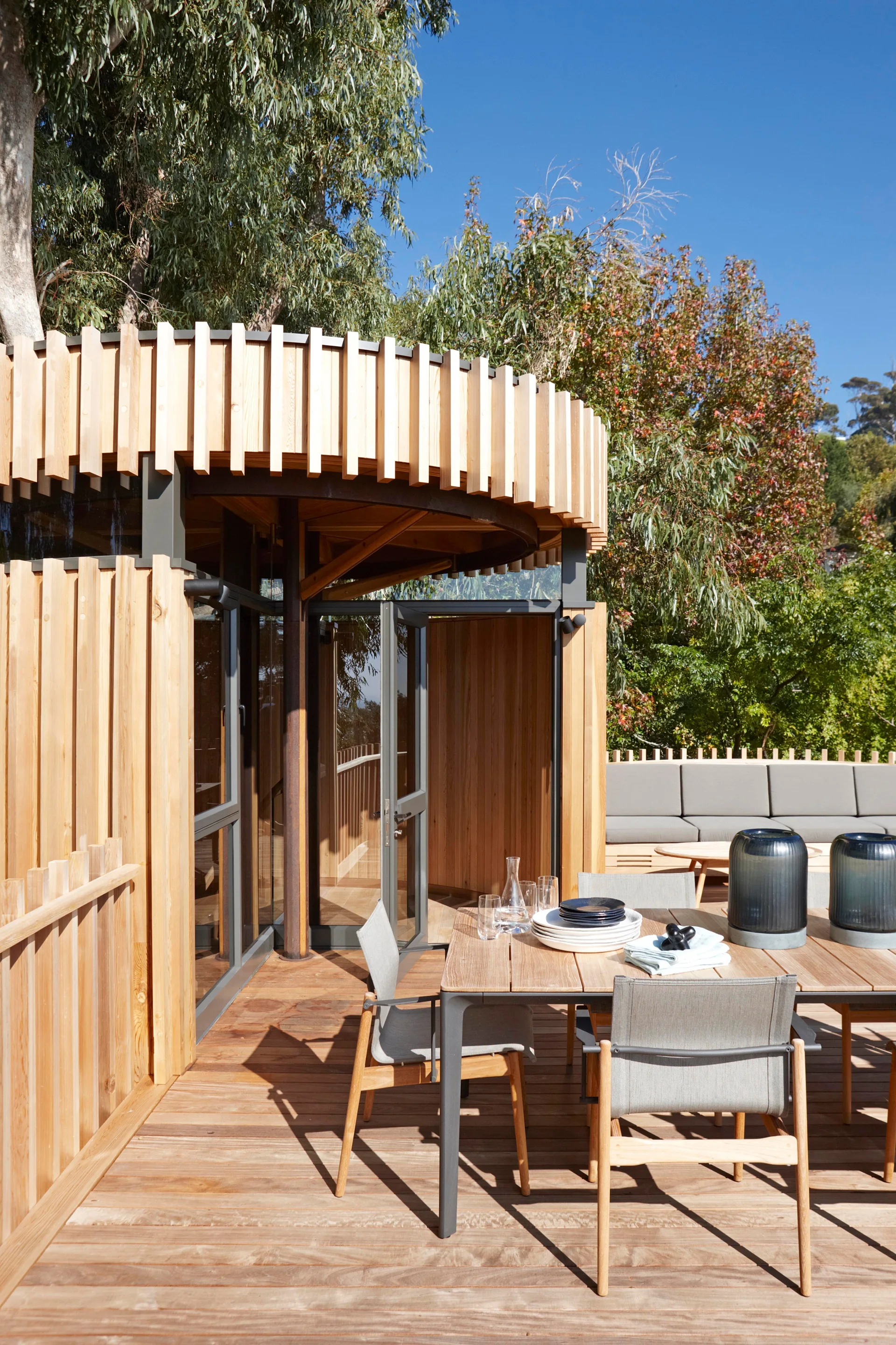 A sunny wooden deck with an outdoor dining table and chairs sits next to a circular timber structure with vertical slats.