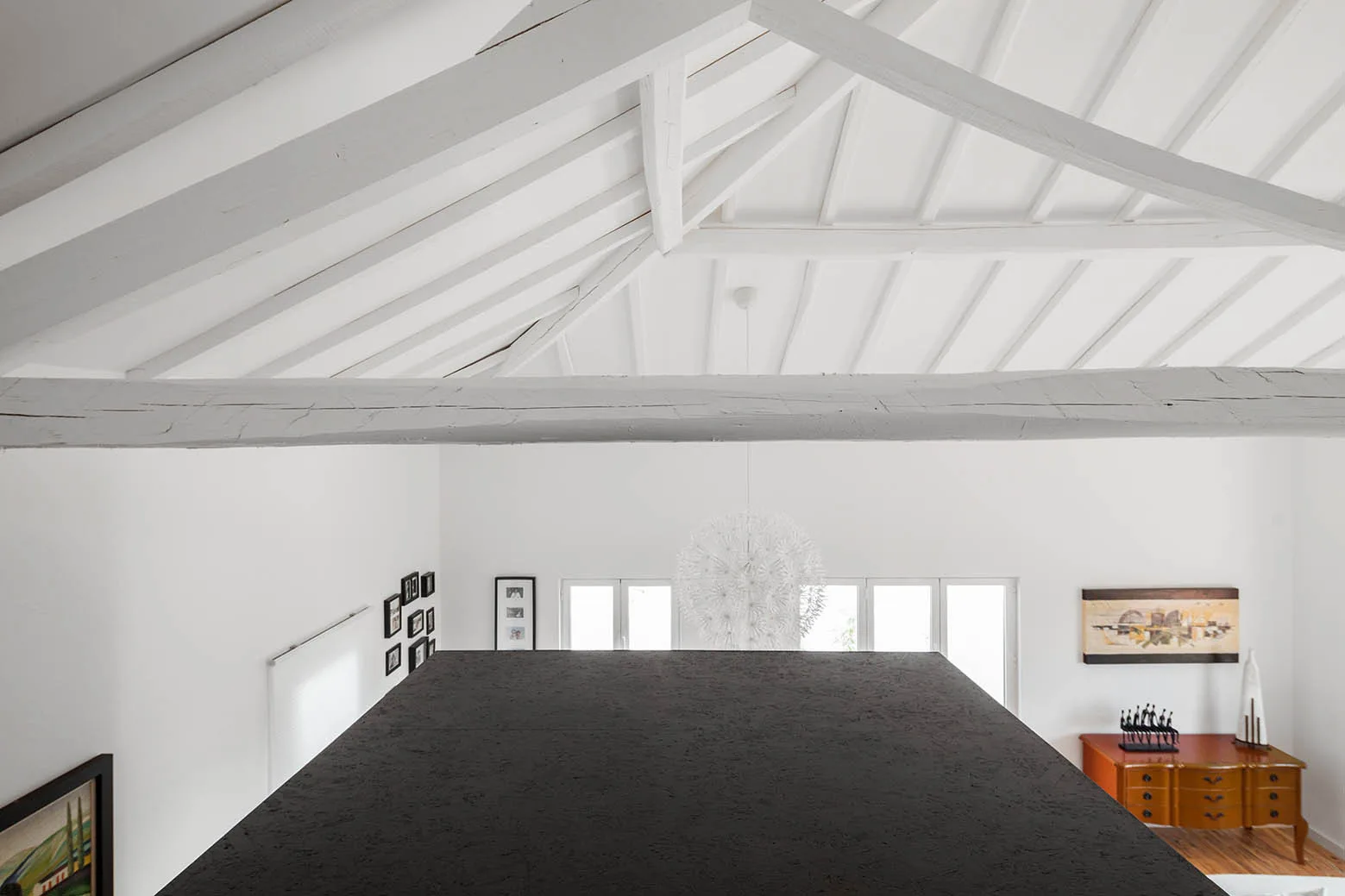 High-angle view from a black mezzanine looking down toward a white living area beneath a pitched ceiling with painted white wooden beams.