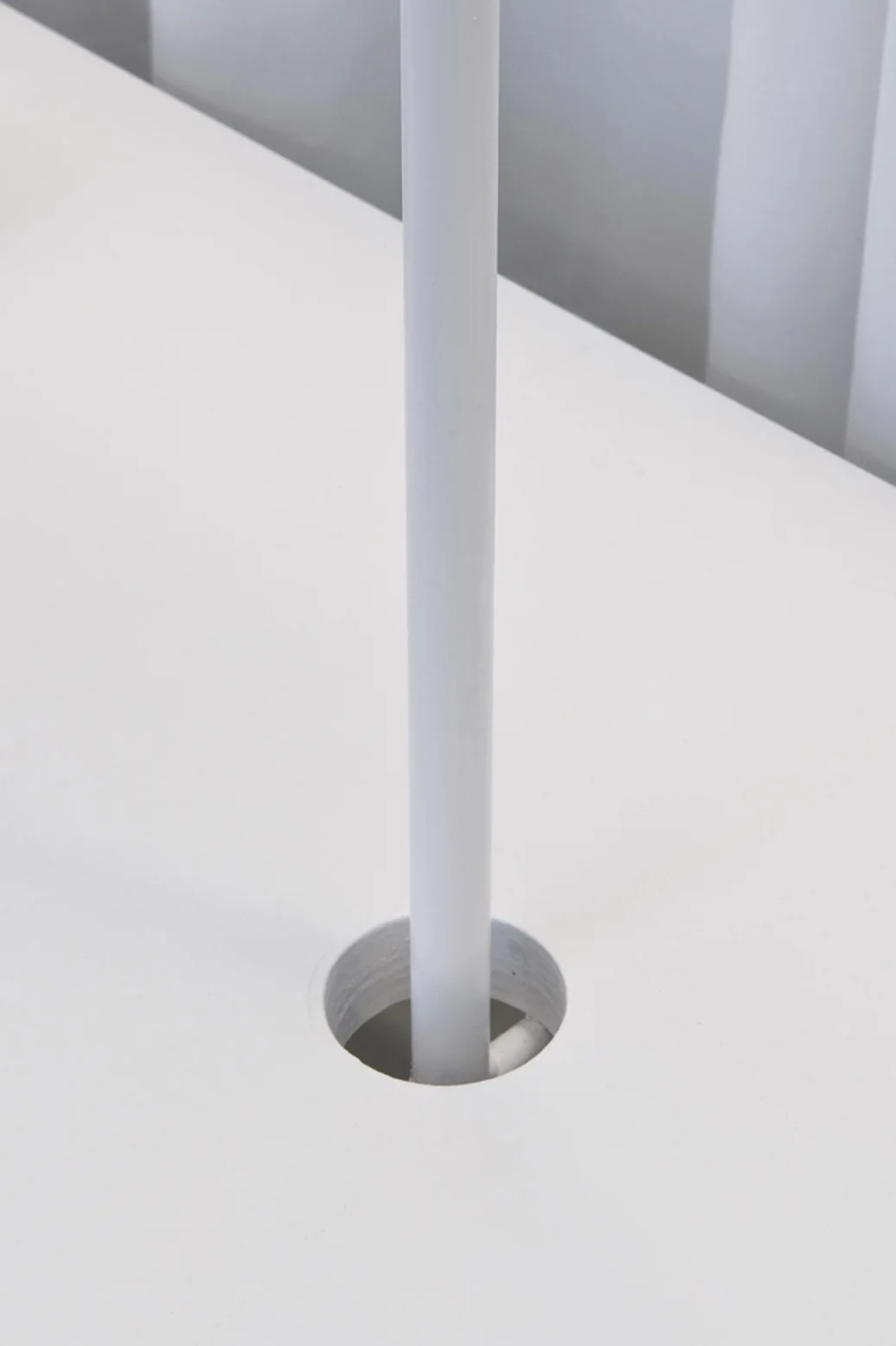 A close-up of a thin white vertical metal pole passing through a circular cutout in a flat matte white desktop.