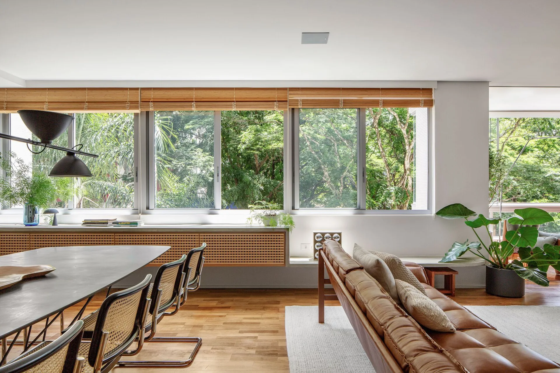 A modern living space features a low tan leather sofa, cane dining chairs, and a long wood lattice console beneath wide windows.