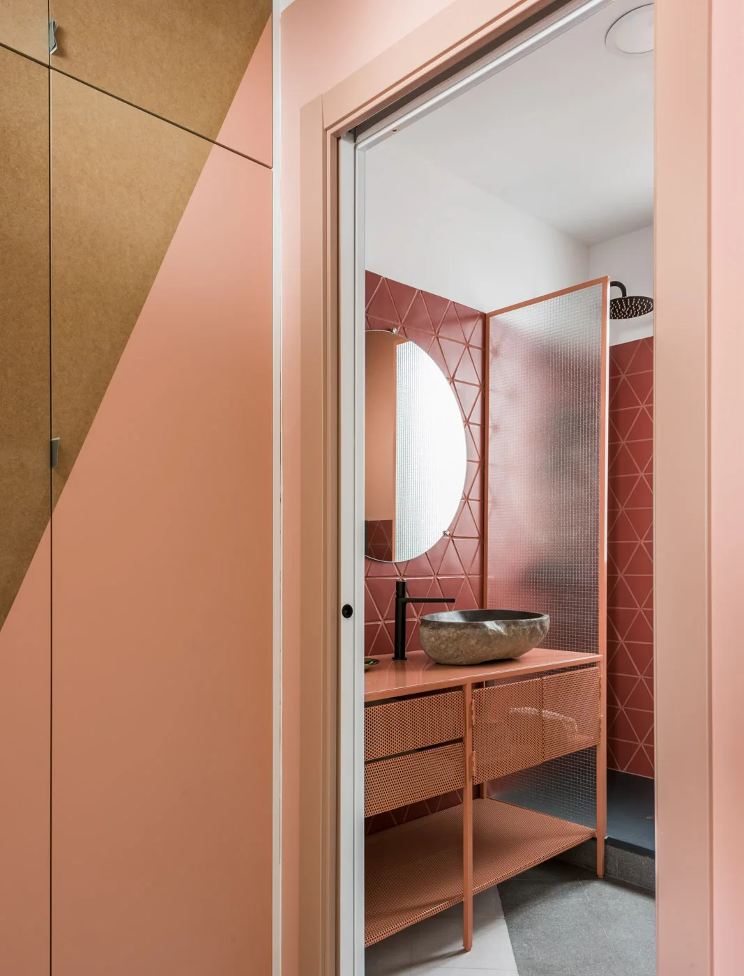 A modern pink bathroom features a rough stone vessel sink atop a perforated metal vanity, backed by triangular terracotta tiles and a round mirror.
