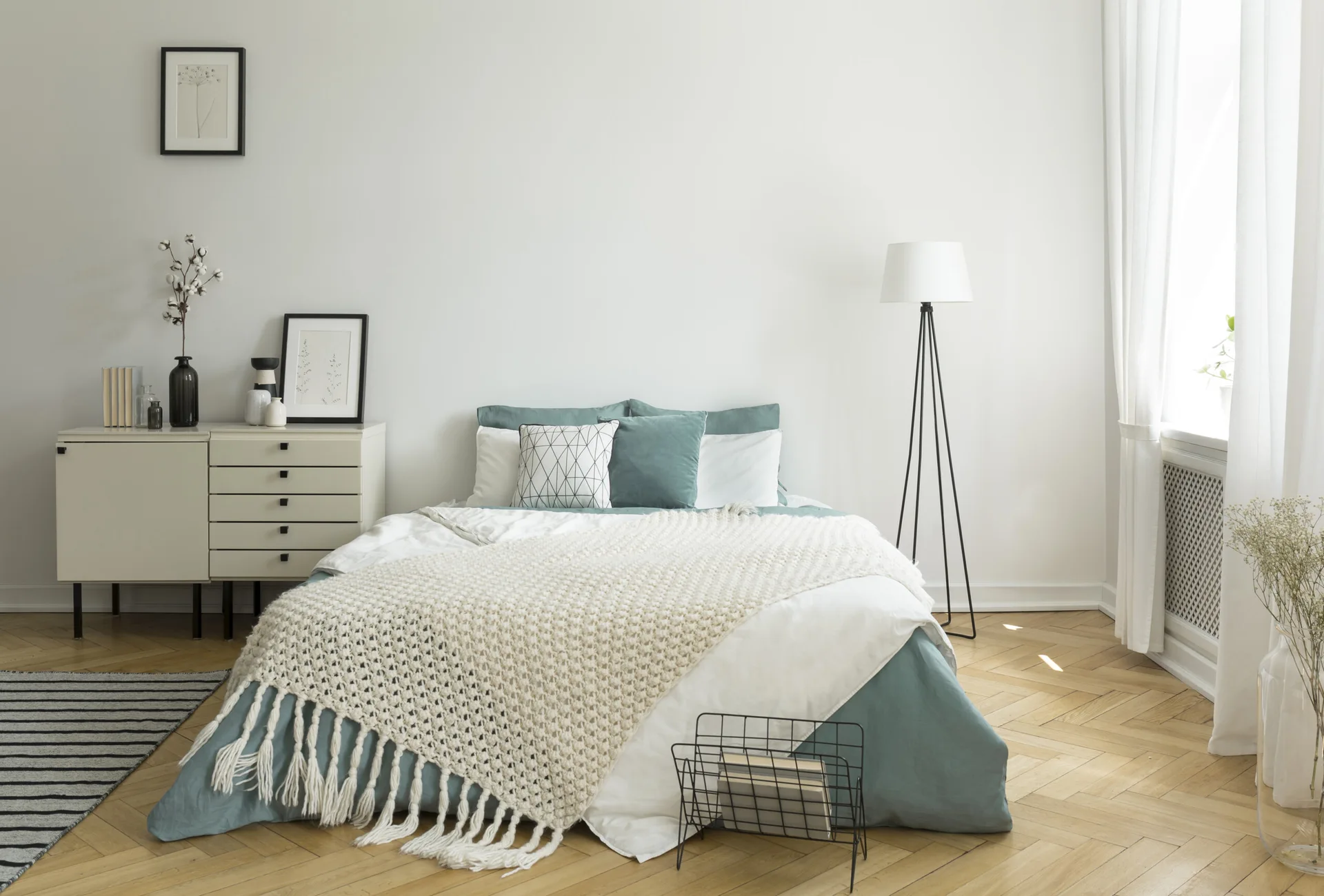 A bright bedroom featuring a bed with sage green linens, a chunky knit throw, a beige dresser, and chevron parquet wooden flooring.