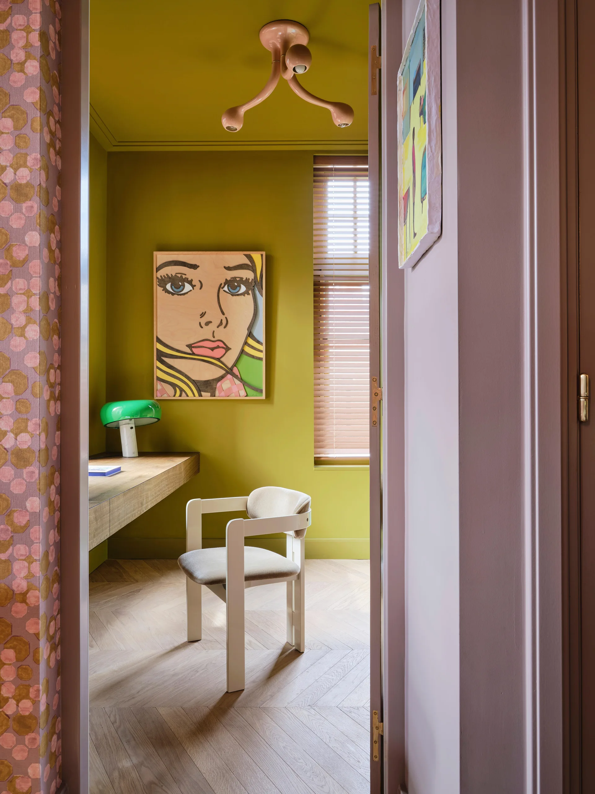 A view through a doorway into a home office with olive green walls, a floating wood desk, a modern white chair, and herringbone flooring.