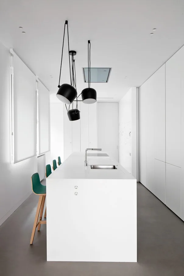 A minimalist white kitchen features a long central island with a sink, a cluster of black pendant lights, and a row of green bar stools.