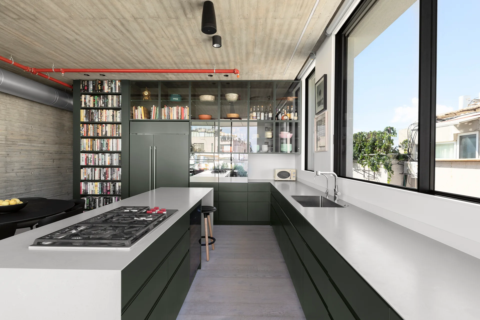 A modern kitchen features dark green matte cabinetry, a light gray countertop island with a gas cooktop, and an exposed board-formed concrete ceiling.