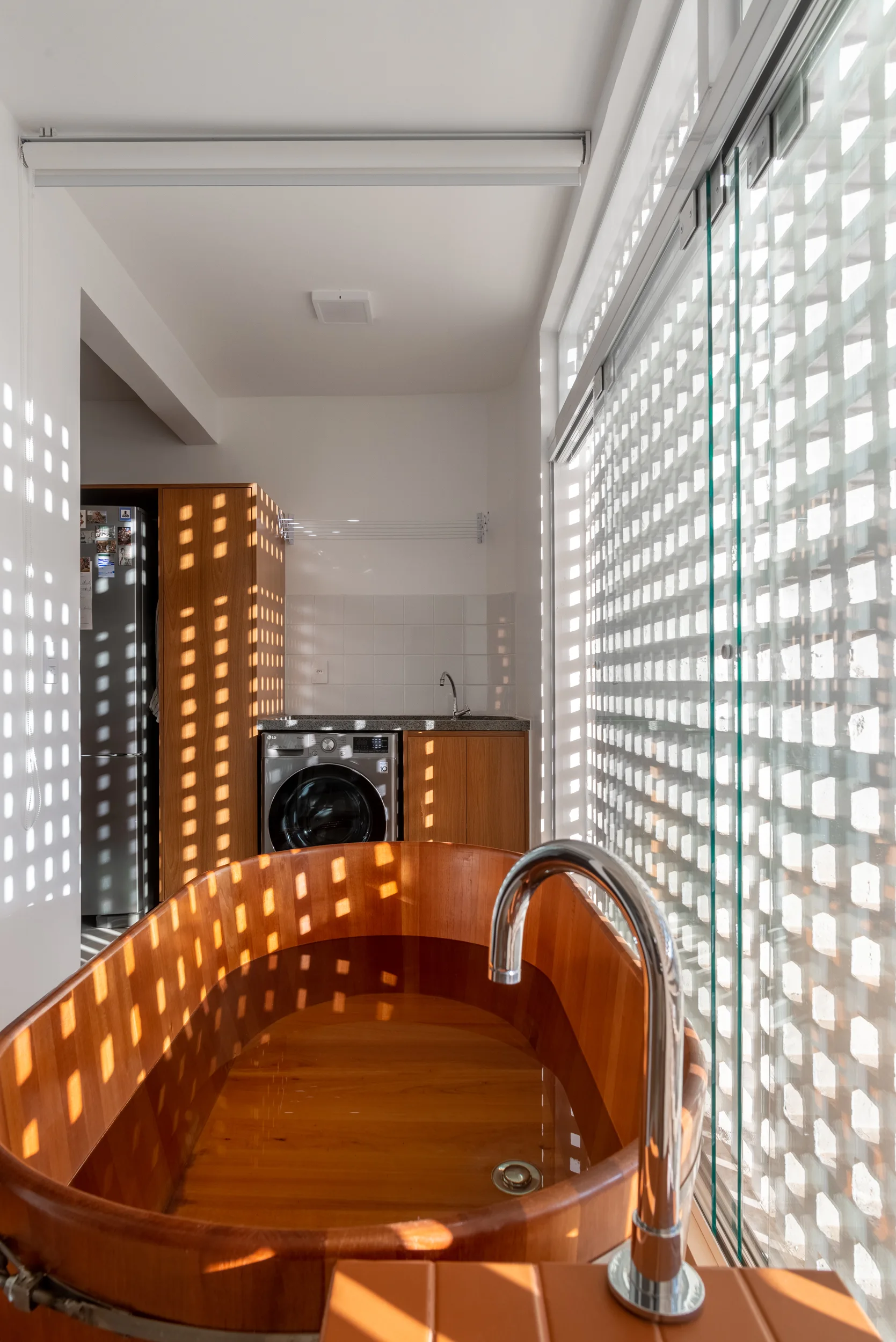 A deep wooden soaking tub stands near a glass wall where an exterior latticed screen casts checkered shadow patterns across the bathroom.