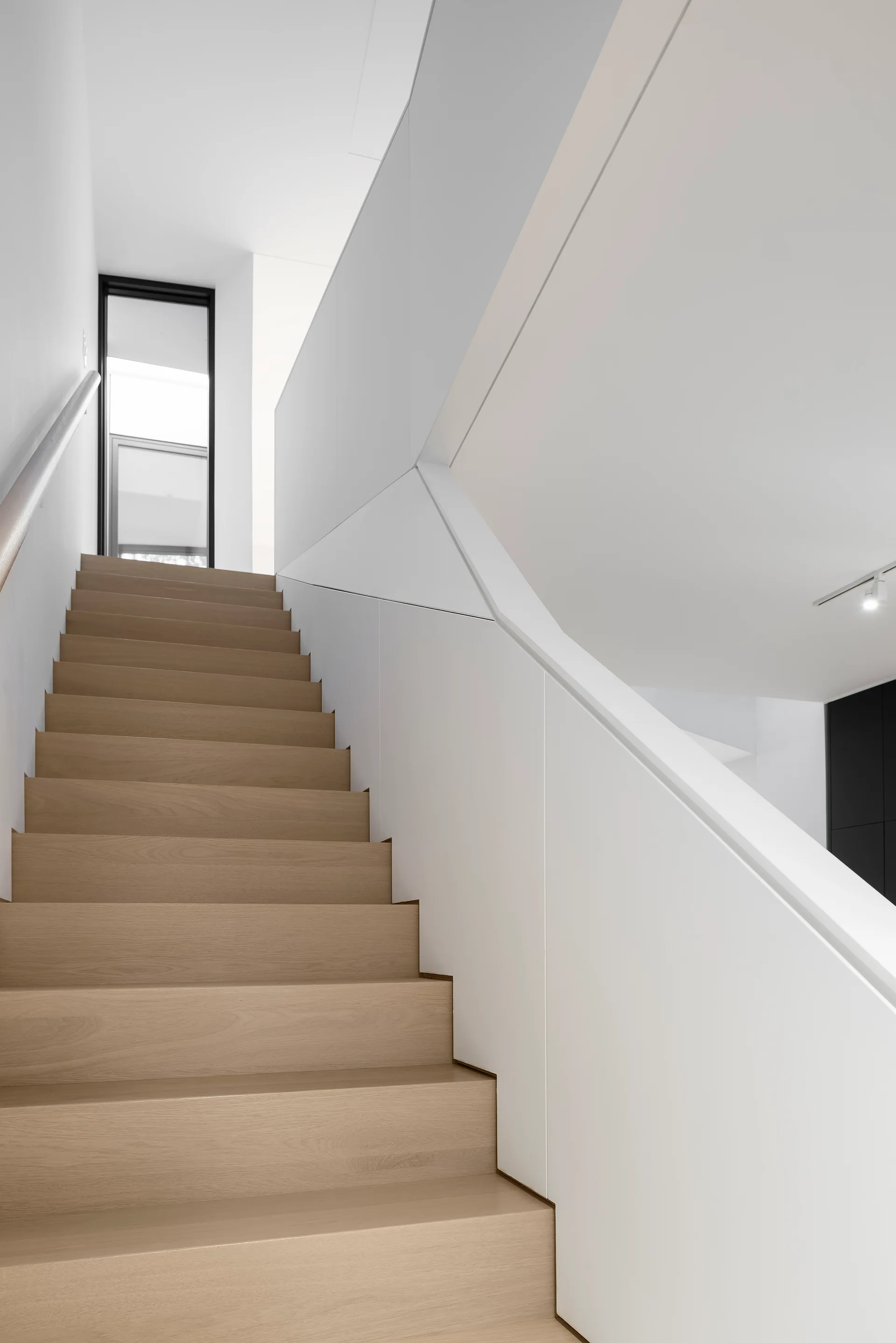 A modern staircase features light wood treads set against solid white walls and a smooth, minimalist white panel balustrade.