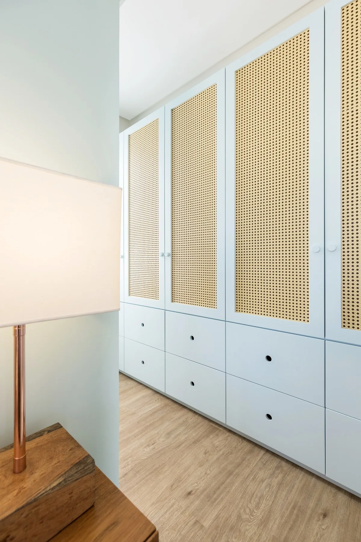 A light blue built-in wardrobe with woven cane door panels stands next to a wooden bedside table with a lighted lamp.