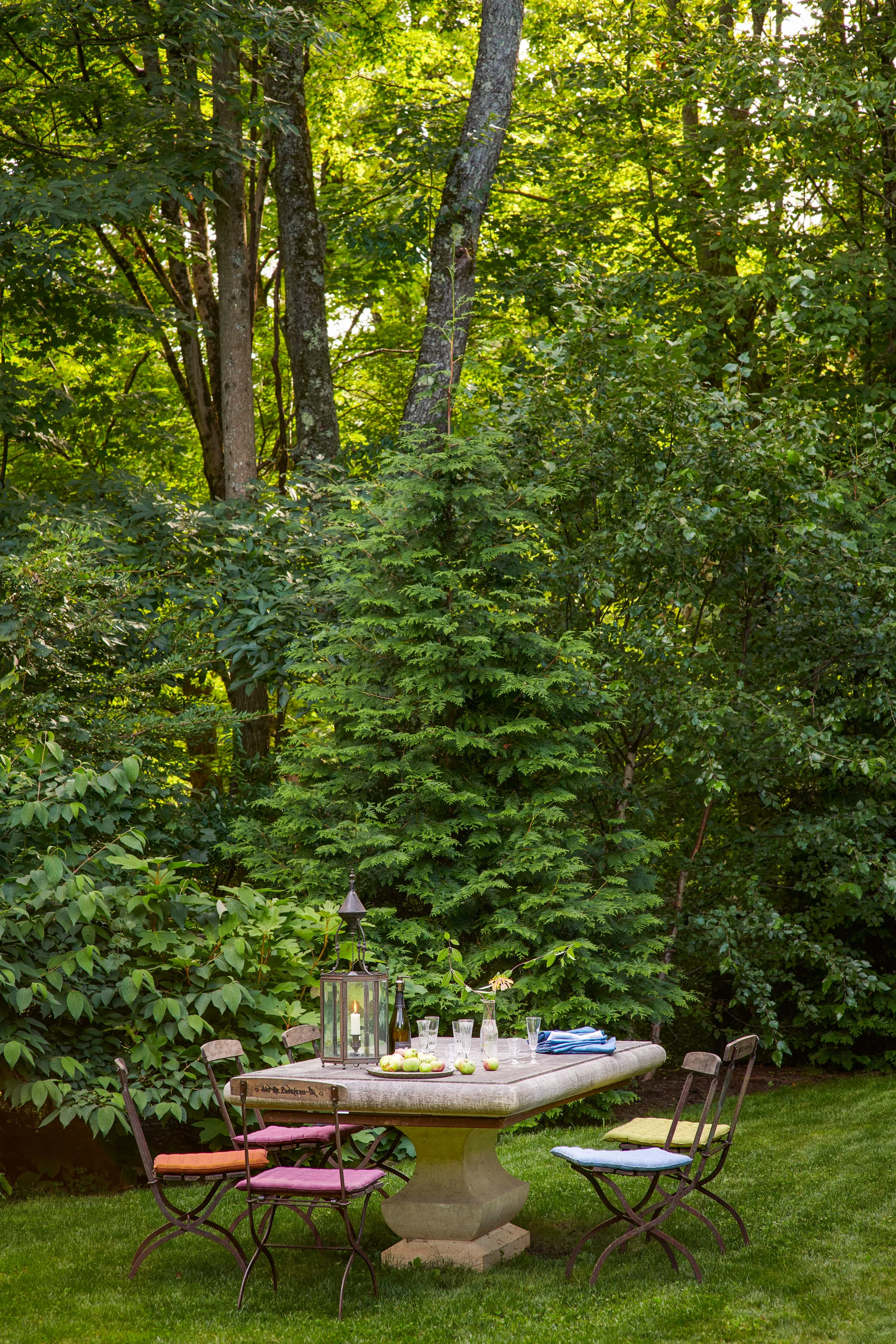 A stone pedestal dining table and metal folding chairs with colorful cushions sit on a green lawn surrounded by dense trees.