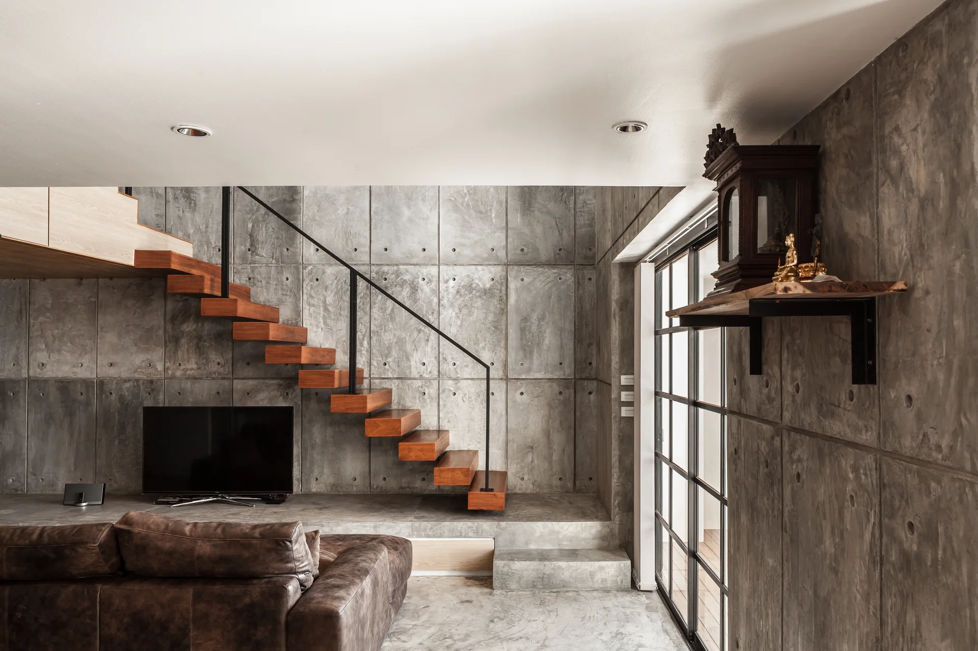 A modern living room features a floating wooden staircase mounted on a grid-patterned concrete panel wall next to a brown leather sofa.