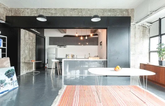 An open-concept dining space and kitchen with exposed concrete walls, a polished grey floor, a striped rug, and an oval white table.